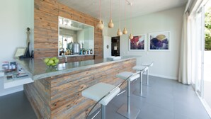 A modern kitchen with a wooden accent wall, a central island with bar stools, pendant lights, and a large window providing natural light. - Sea Five Boutique Hotel (Cape Town)