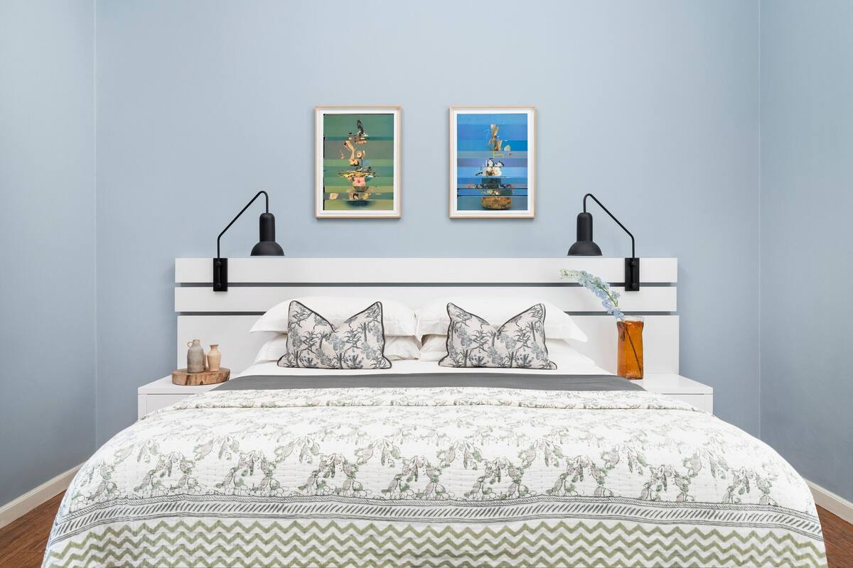 A bedroom with a bed, two wall-mounted lamps, a nightstand, a vase, and two framed artworks.