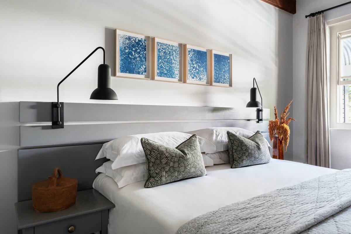 A neatly arranged bedroom with a bed, bedside lamps, a nightstand, and a wall-mounted shelf with framed artwork.