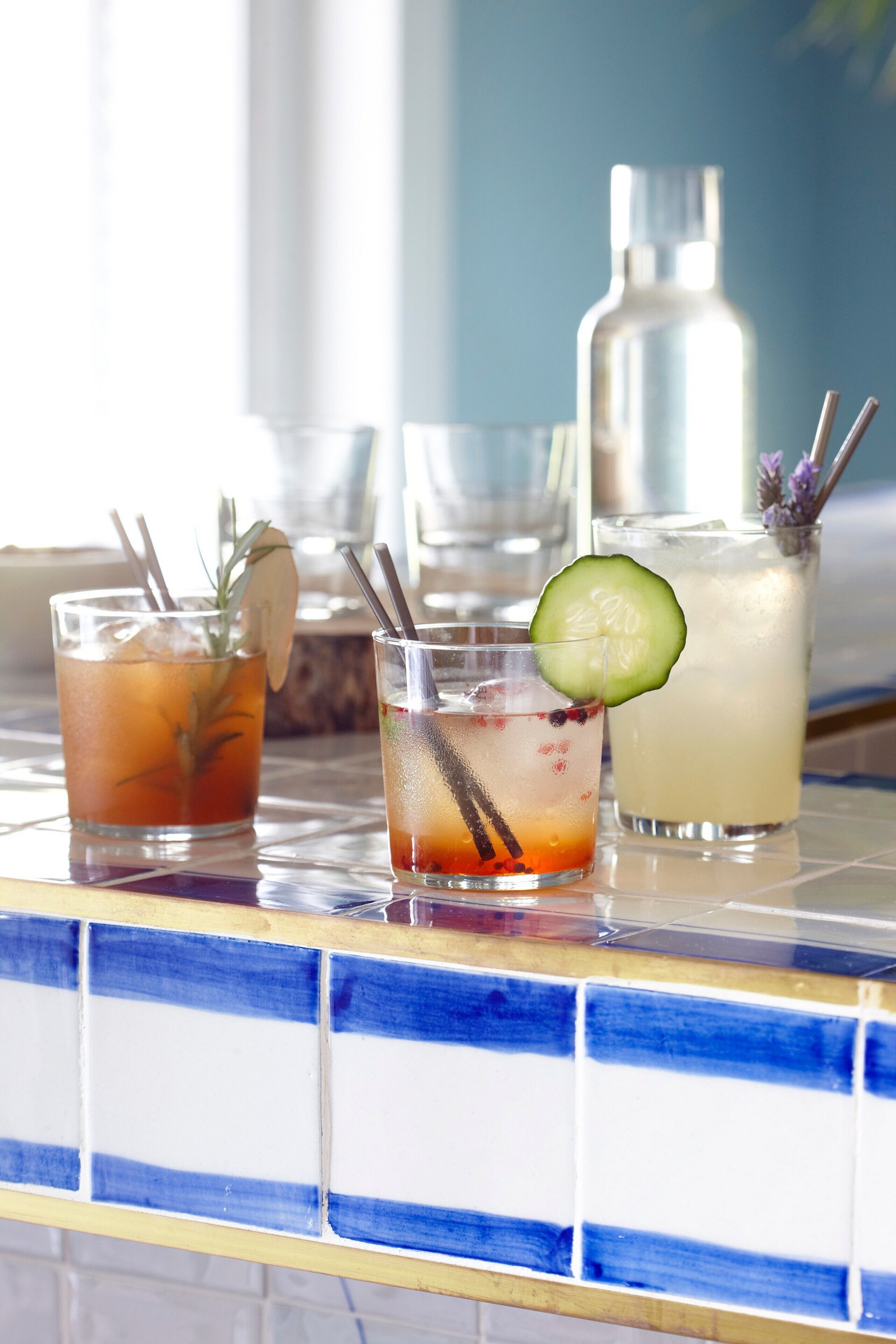 Three glasses of beverages on a tiled counter with a bottle in the background.