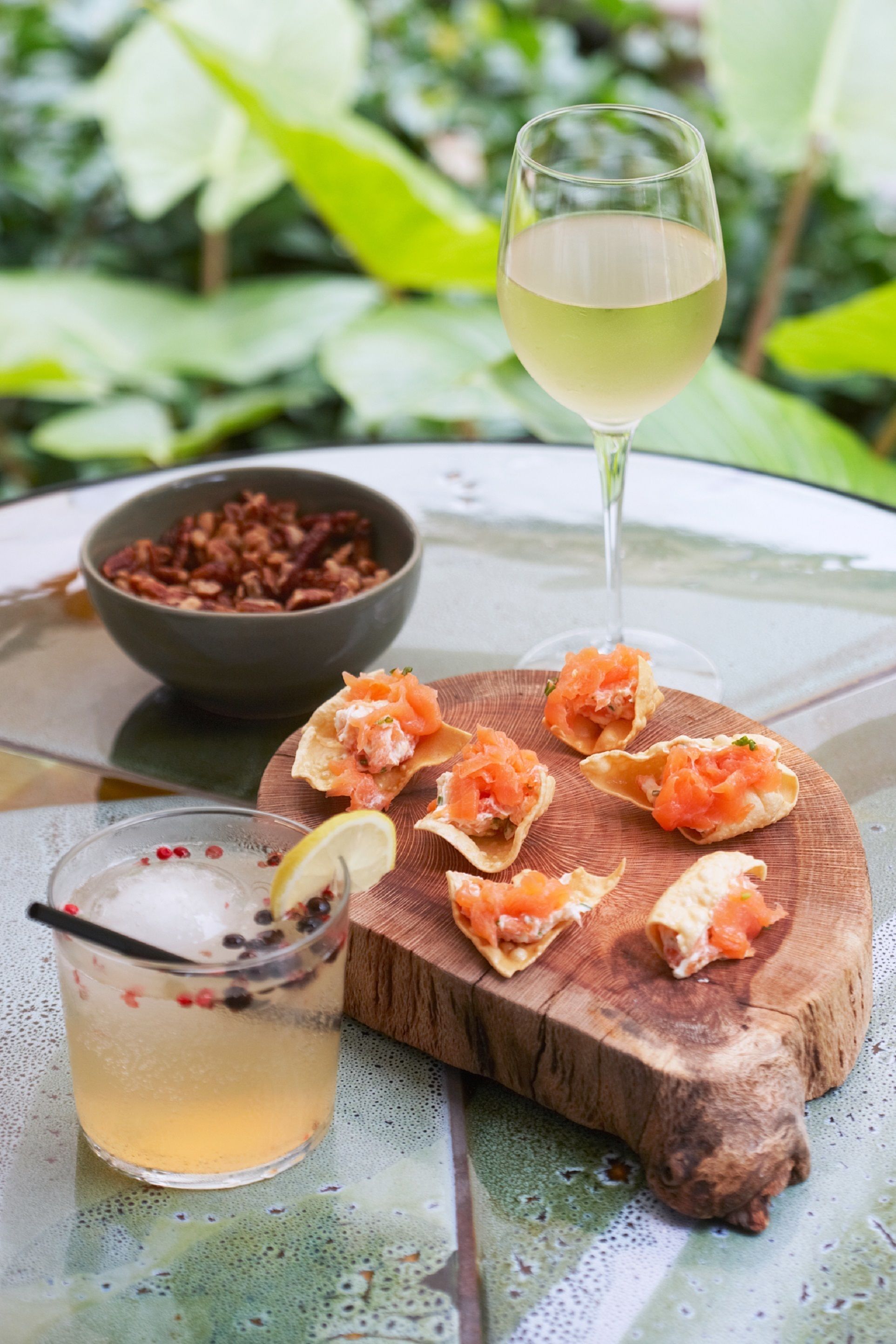 A table with a glass of white wine, a bowl of nuts, and a wooden board with salmon appetizers.