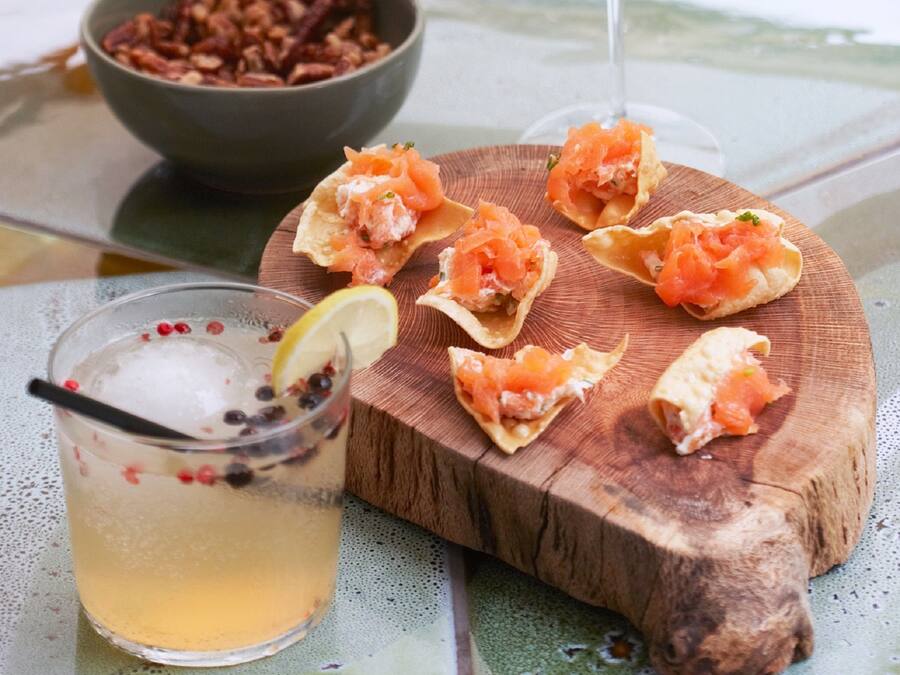 A table with a glass of white wine, a bowl of nuts, and a wooden board with salmon appetizers.