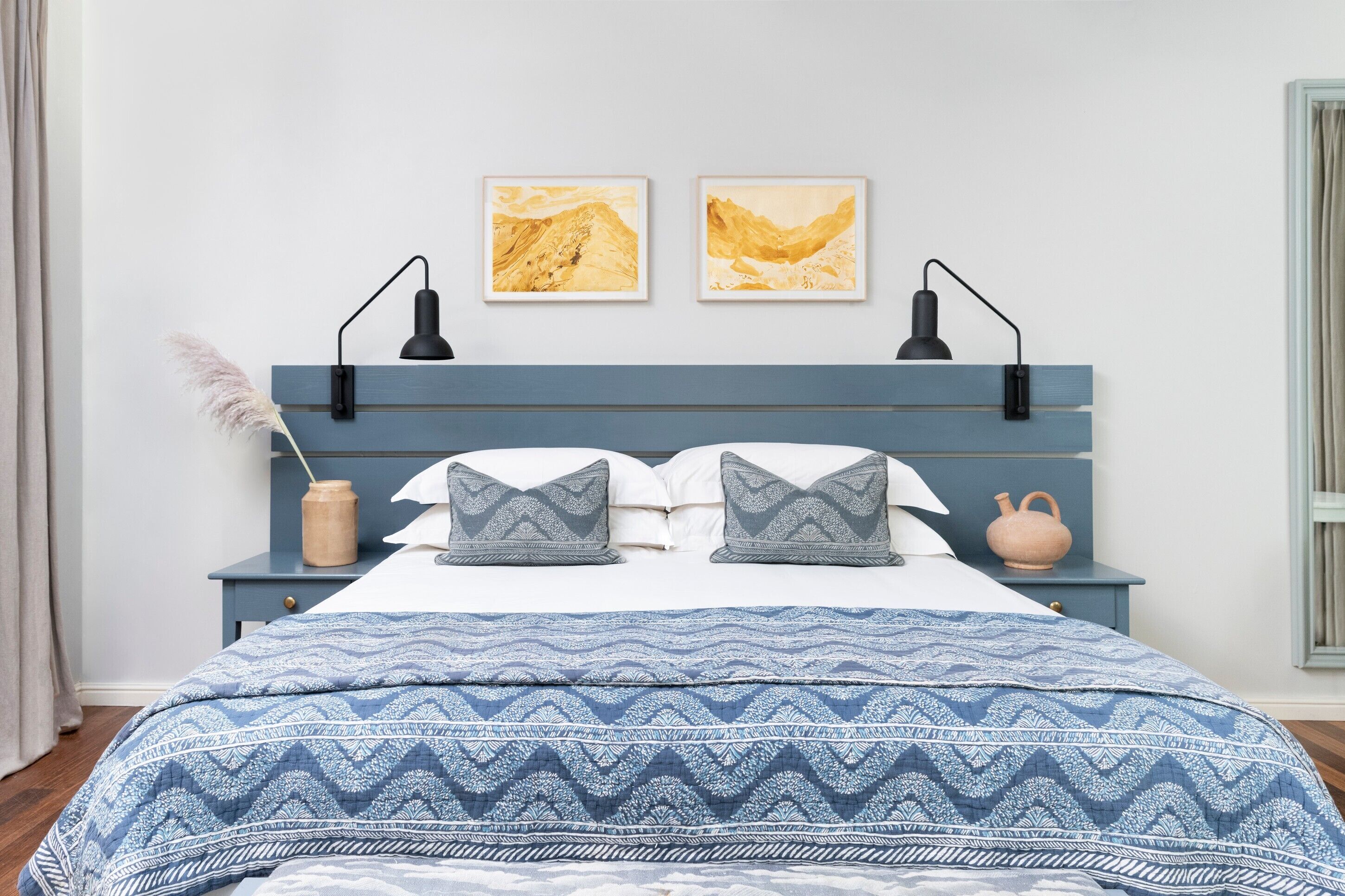 Un dormitorio con cama, dos mesitas de noche, una lámpara de pared, un jarrón con flores secas y dos cuadros en la pared.