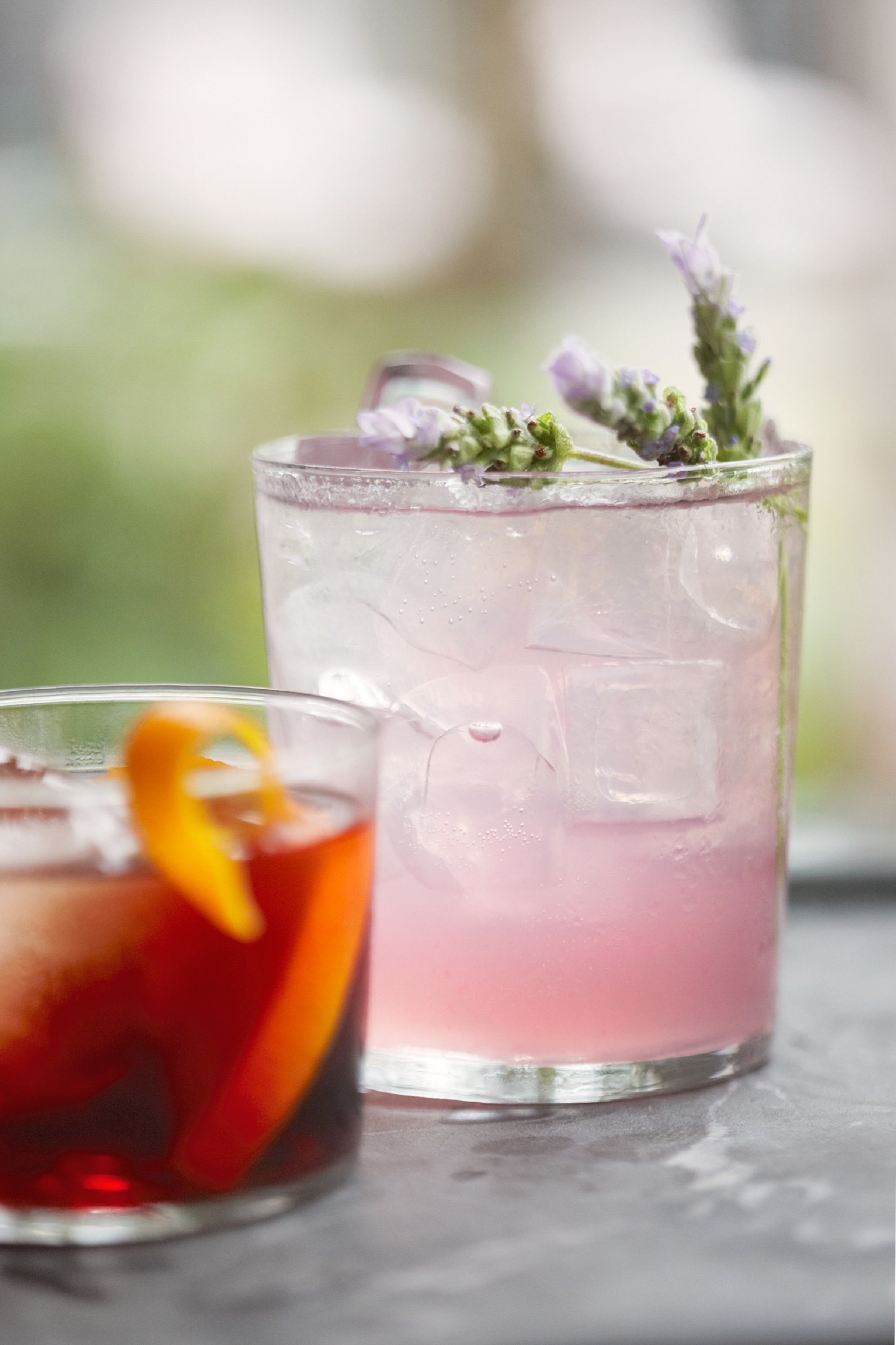 Two glasses of iced drinks with garnishes, one with a red drink and the other with a pink drink, on a table.