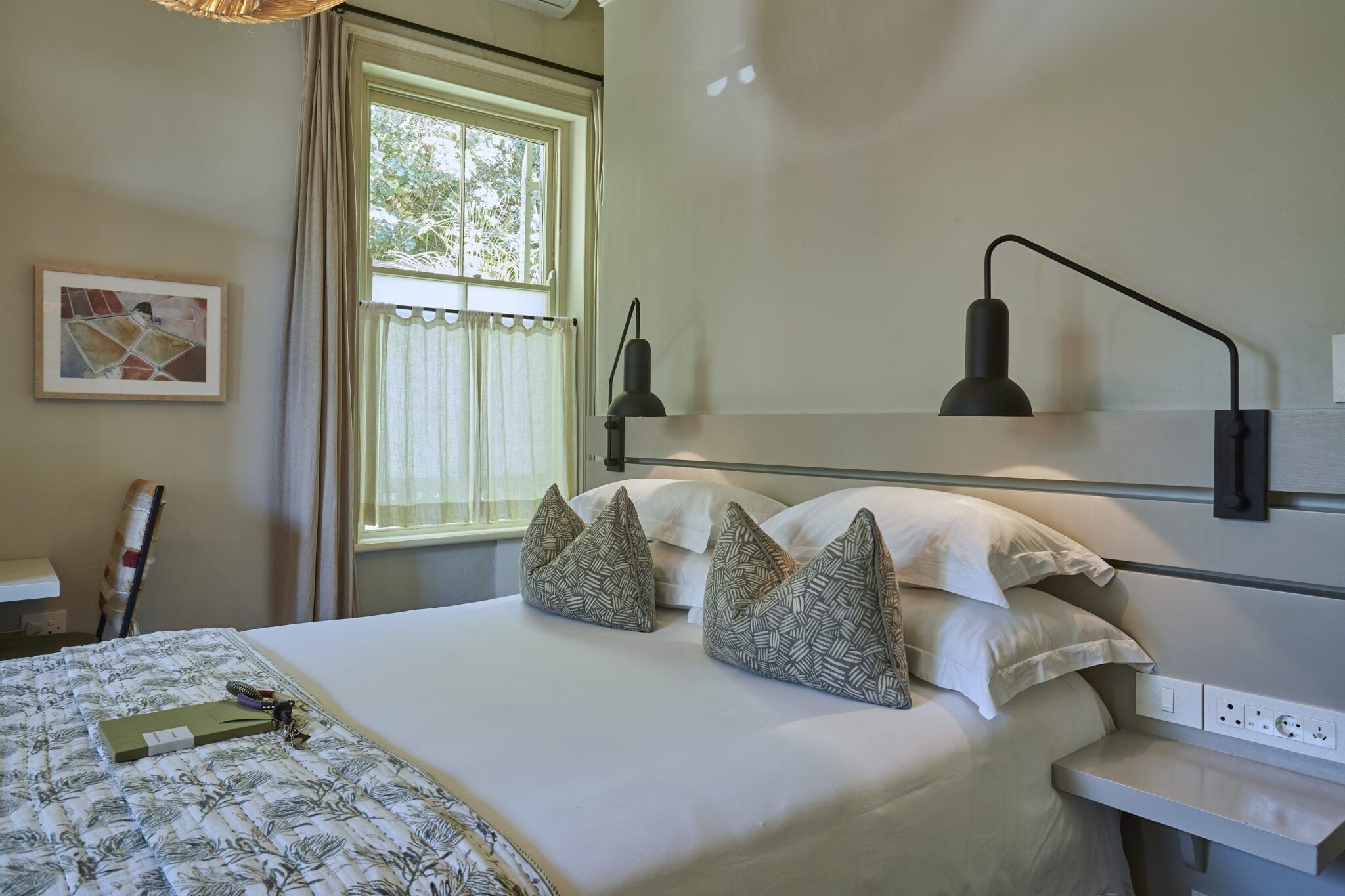 Cama bem arrumada com almofadas estampadas, mesa de cabeceira com abajur e janela com cortinas.