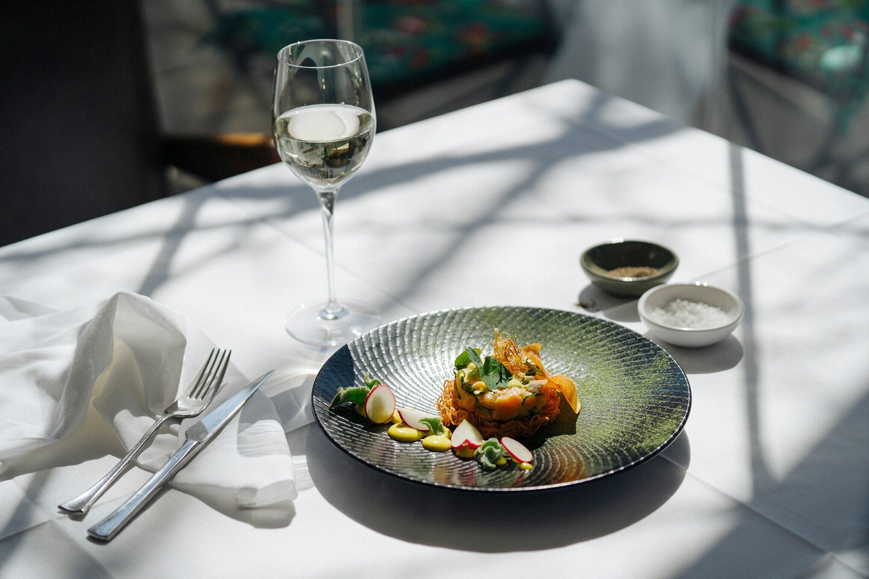 Une table à manger avec une assiette de nourriture, un verre de vin blanc et un bol de sel.