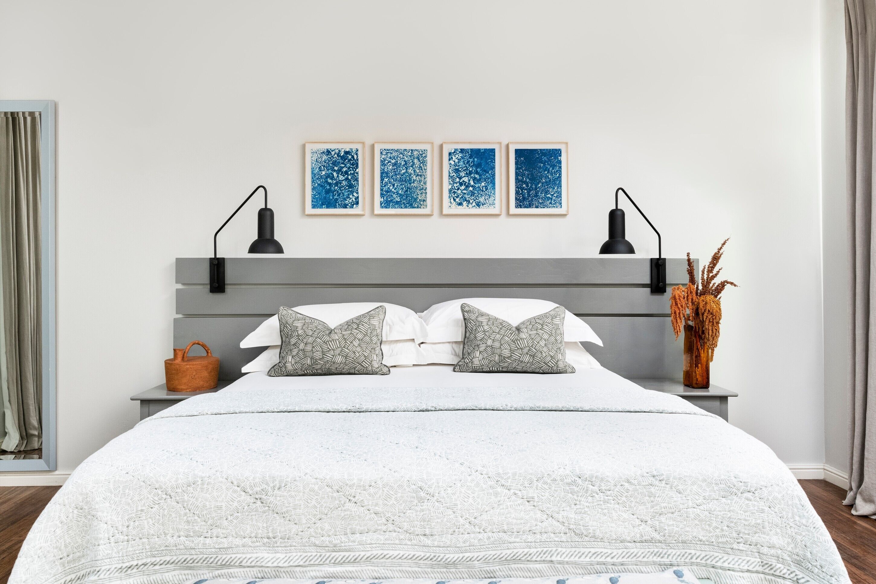 A neatly made bed with a white duvet, two pillows, and a grey headboard. There are two bedside tables with black lamps, a small orange basket, and a vase with dried flowers. Three blue artwork pieces are hung on the wall above the bed.