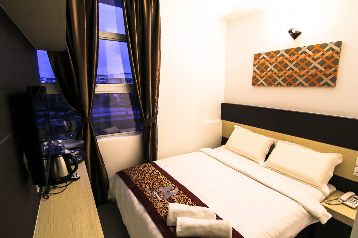 A hotel room with a bed, a desk with a kettle, a telephone, and a window with curtains.