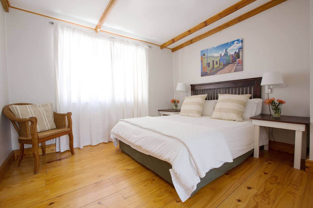 Quarto com piso de madeira, cama com roupa de cama branca, uma cadeira de vime, mesa de cabeceira com abajur e um quadro na parede.