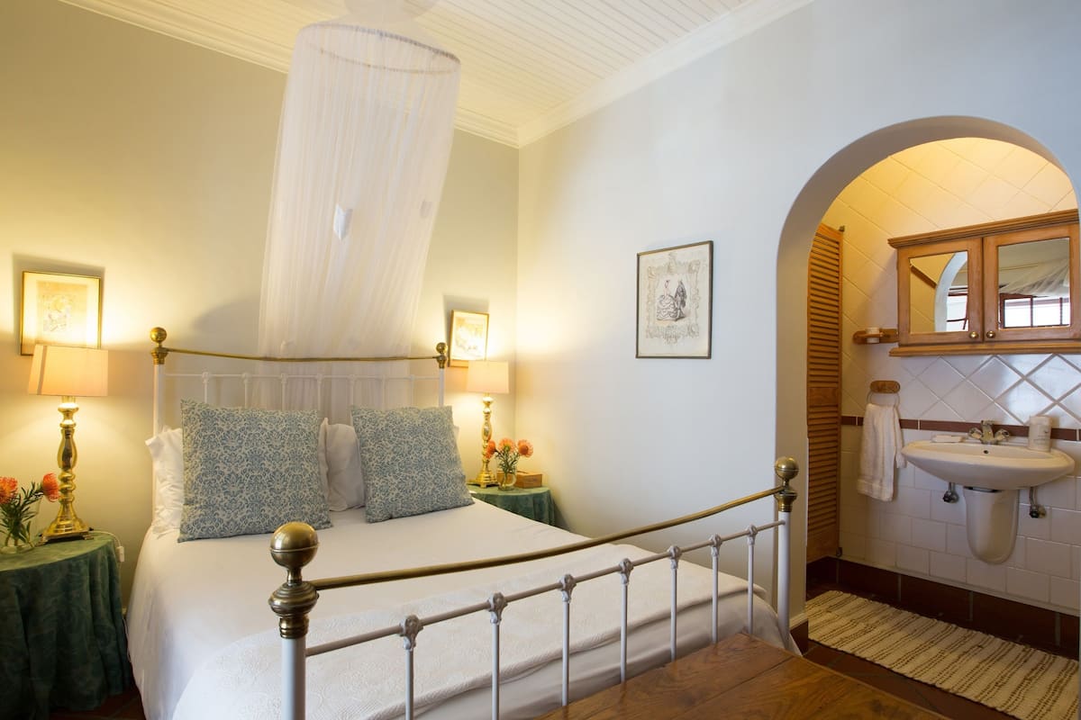 Quarto com cama de dossel, mesa de cabeceira com abajur e banheiro com pia e espelho.