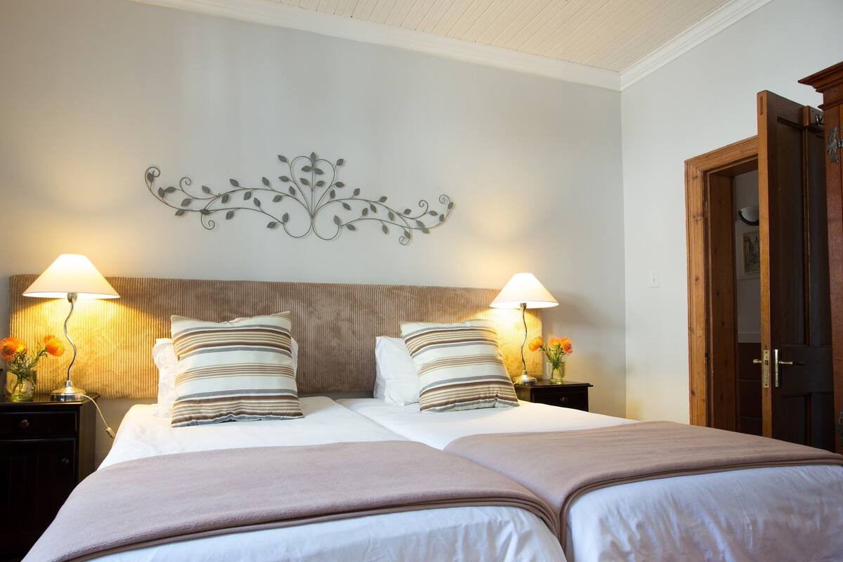 Uma cama de casal com duas mesas de cabeceira, cada uma com uma luminária e um vaso de flores. Uma peça decorativa está pendurada acima da cama.