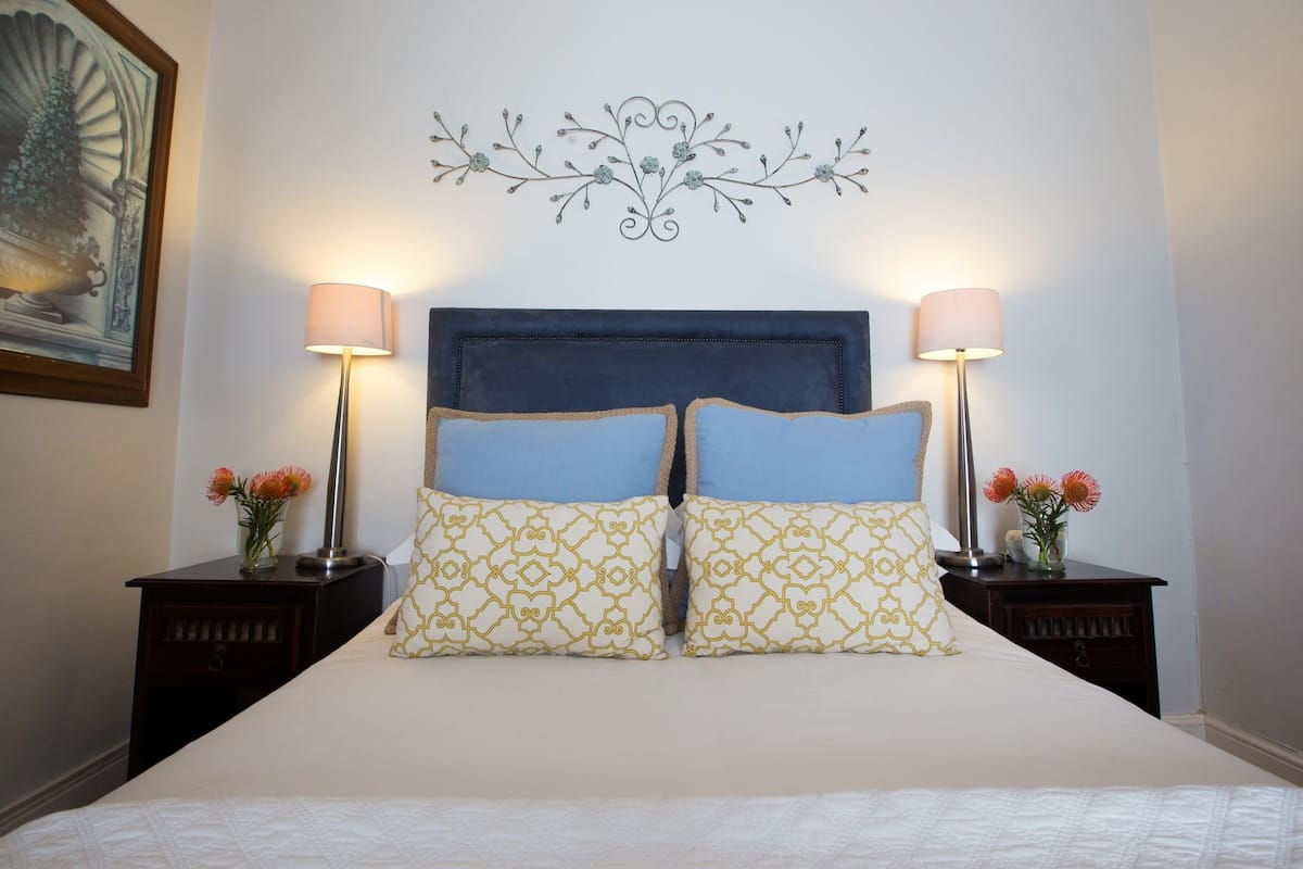 Cama bem arrumada com cabeceira azul, duas mesas de cabeceira com abajures e um objeto decorativo na parede acima da cama.