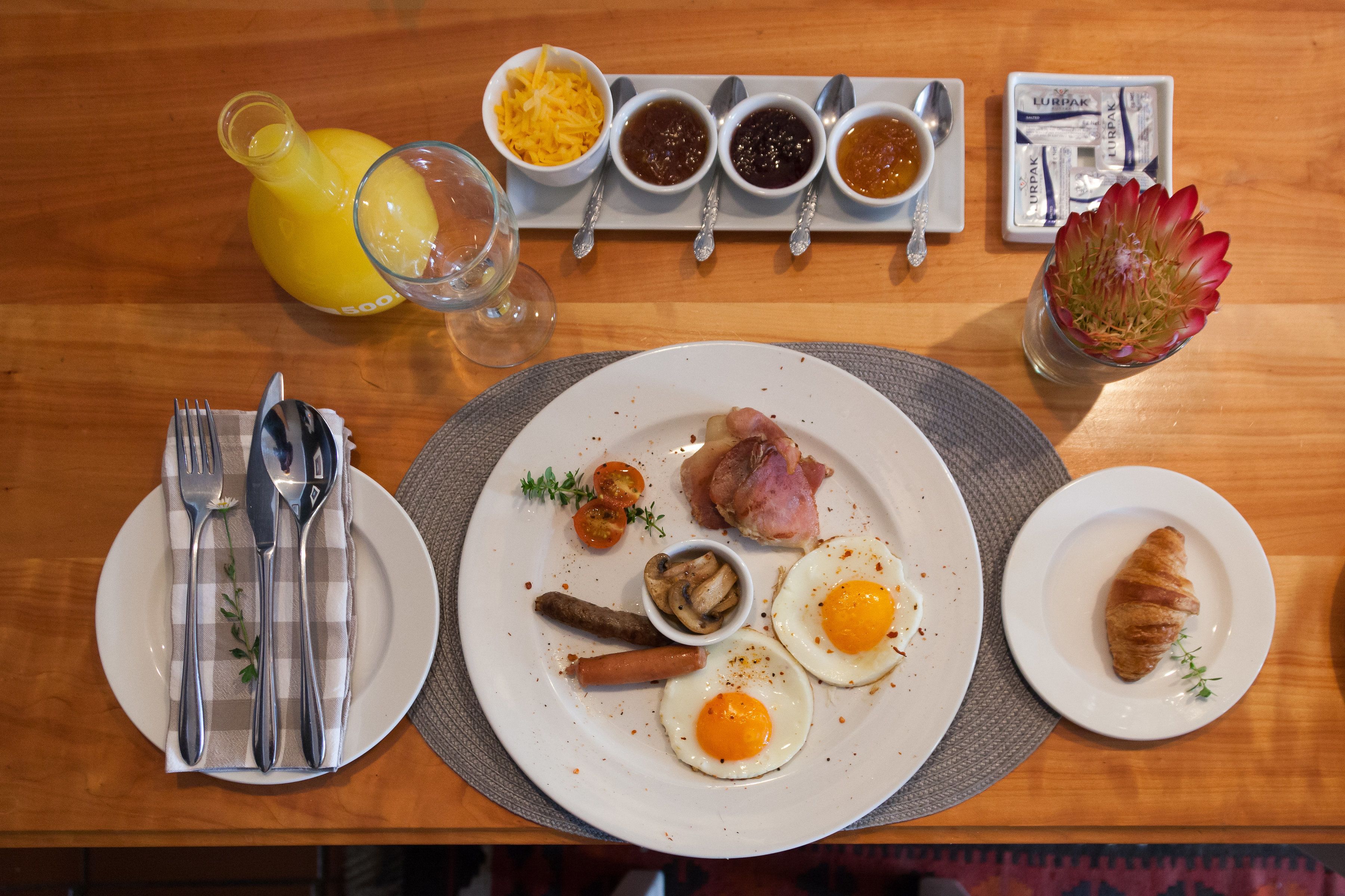 A breakfast table with plates of eggs, ham, sausages, and a croissant, accompanied by condiments and a glass of orange juice.