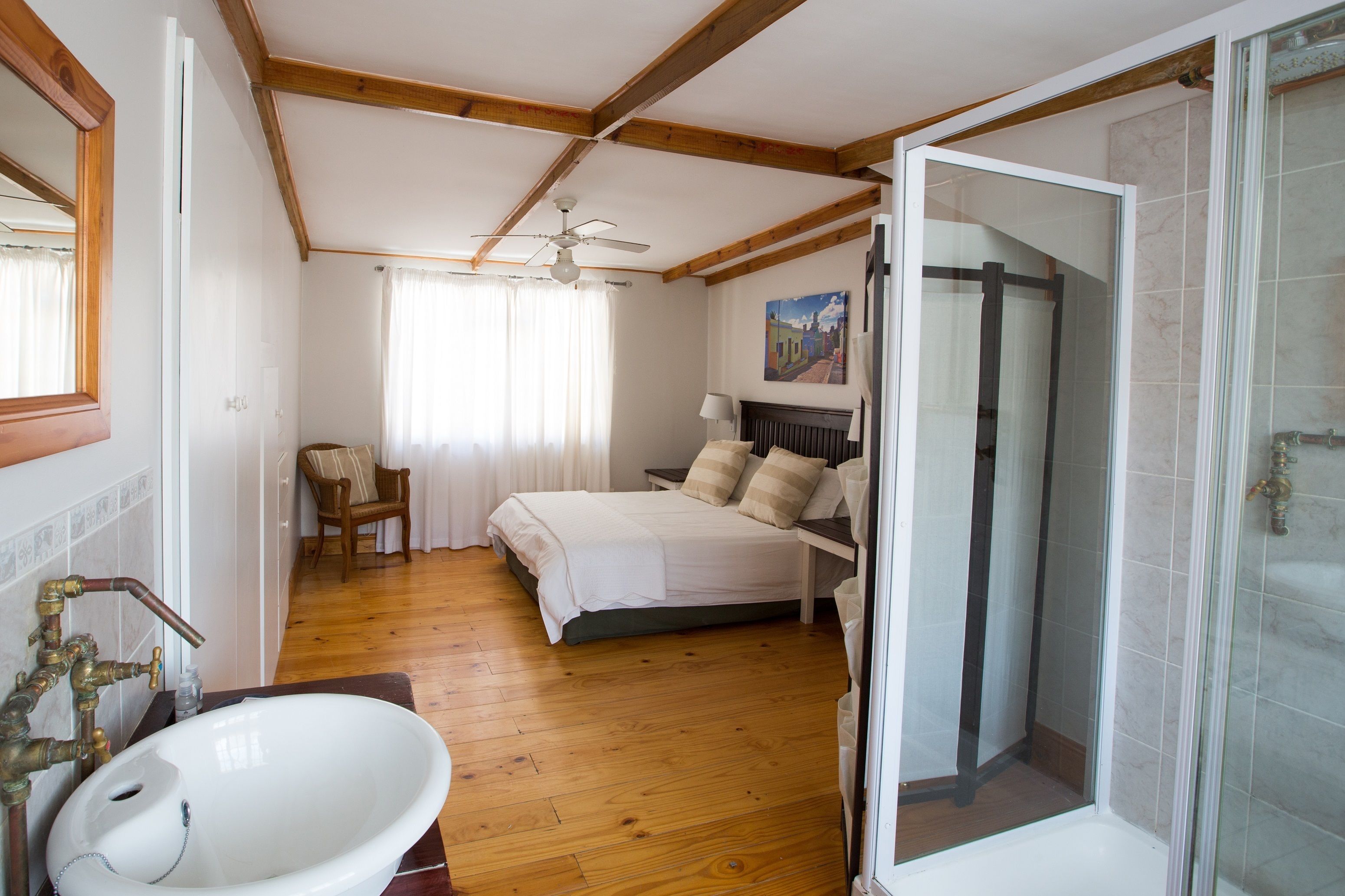 A bedroom with a bed, a bedside table, a chair, a window with curtains, a shower area, and a bathroom with a sink and mirror.