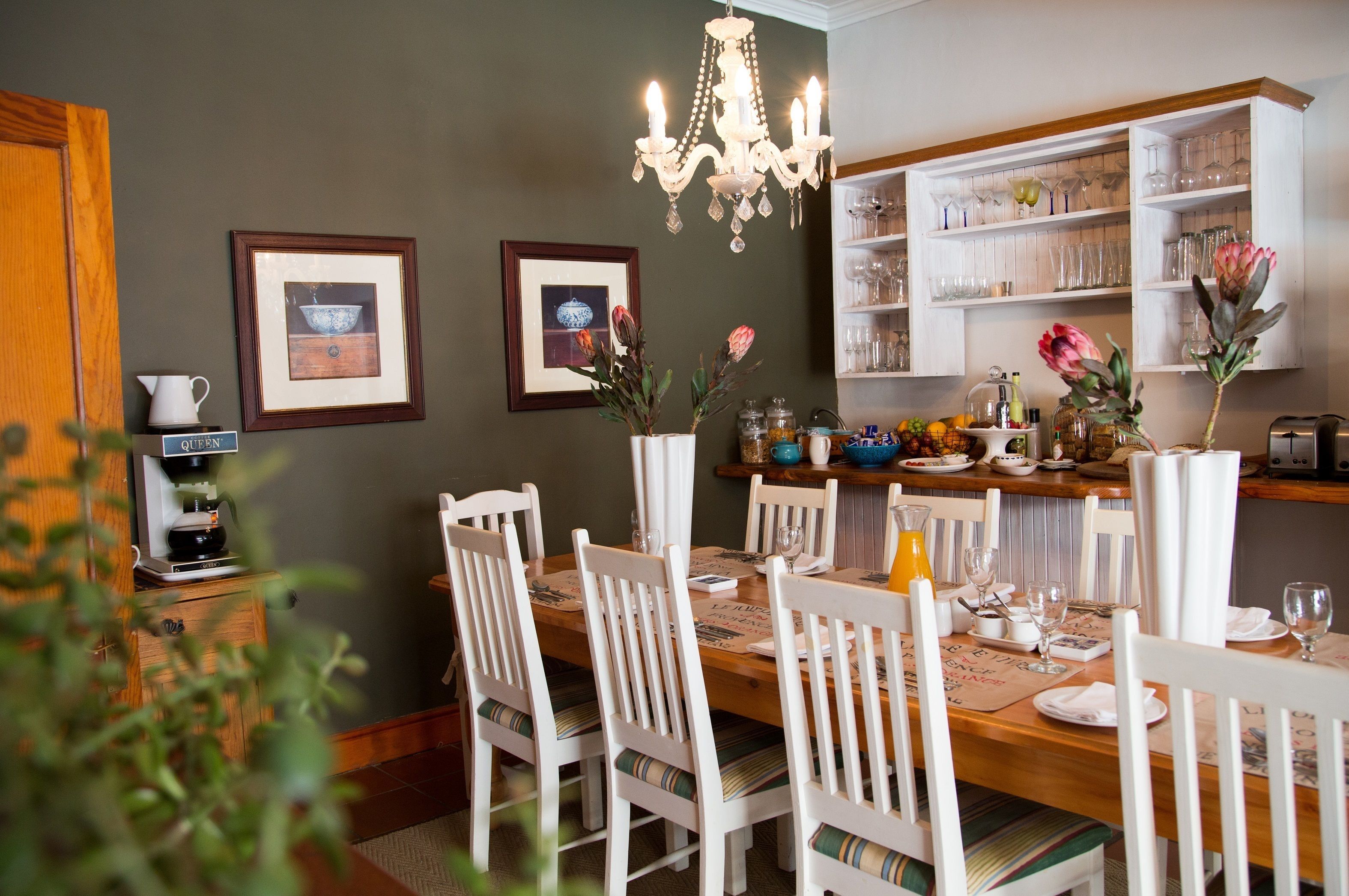A dining area with a wooden table, white chairs, a chandelier, framed pictures on the wall, and a kitchen area with shelves and jars.