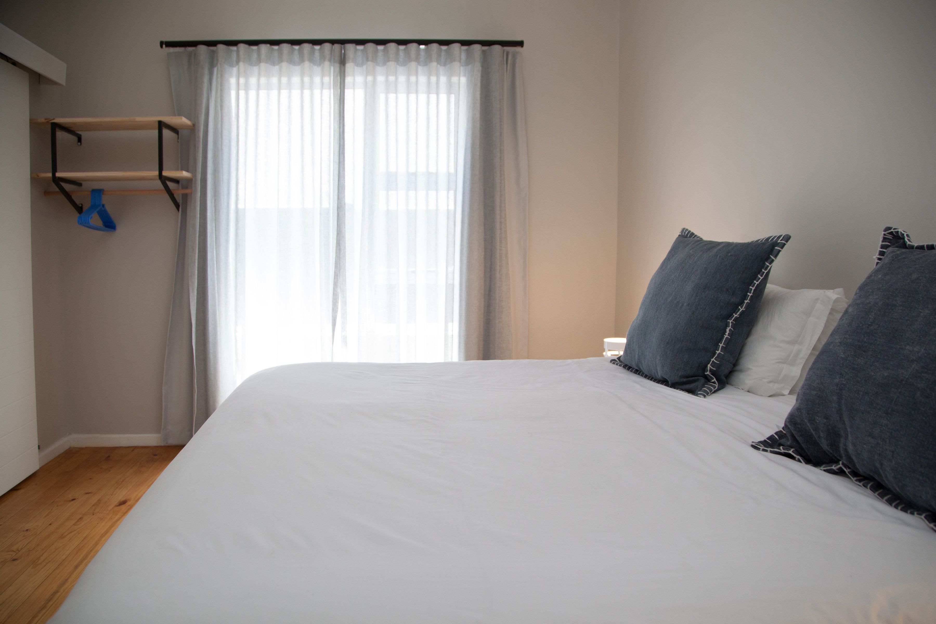 Un dormitorio con una cama grande, suelo de madera, ventana con cortinas traslúcidas y un estante en la pared con un objeto azul.