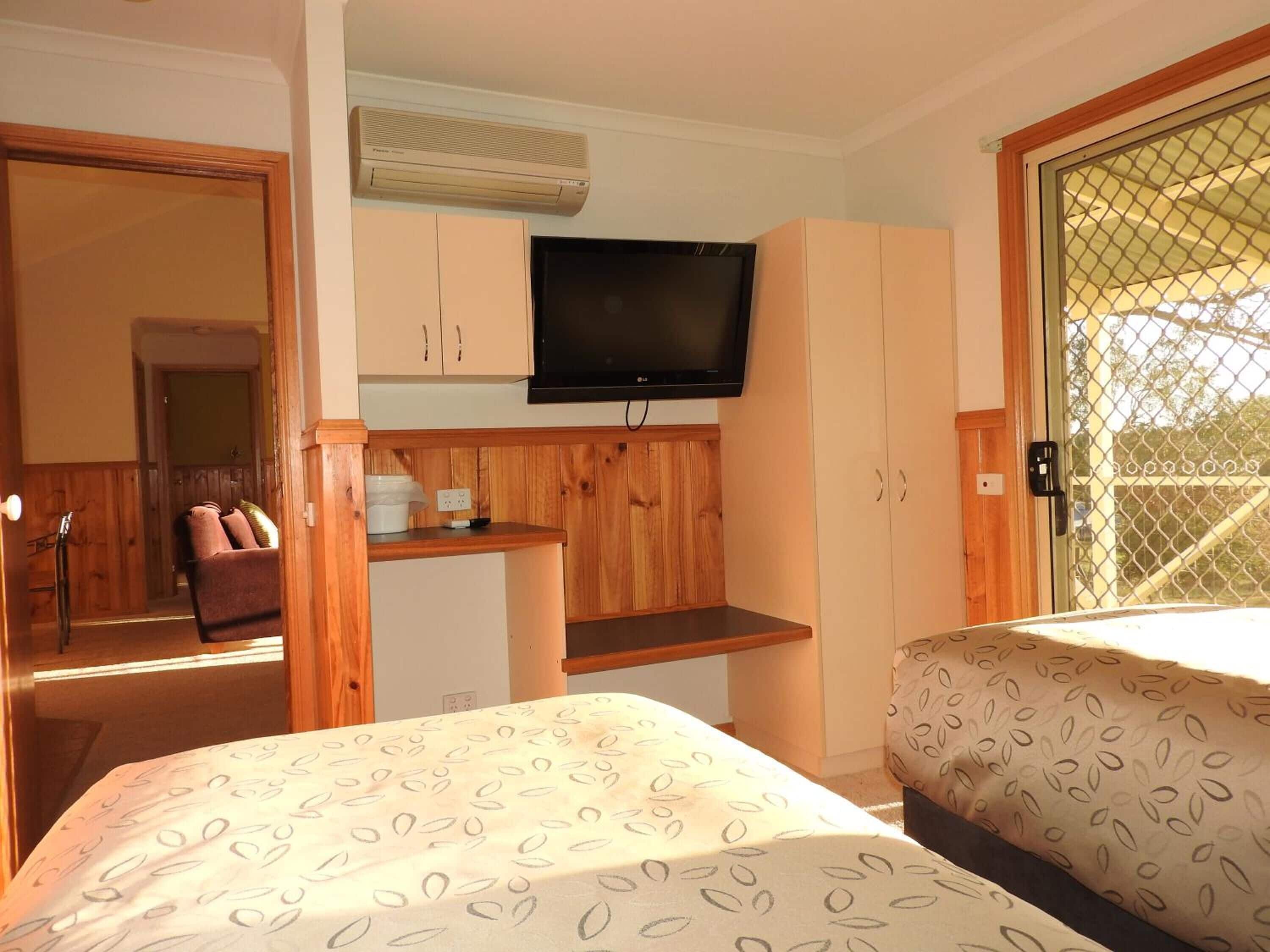 Una habitación con cama, televisor montado en la pared, armarios de madera y ventana con mosquitero.