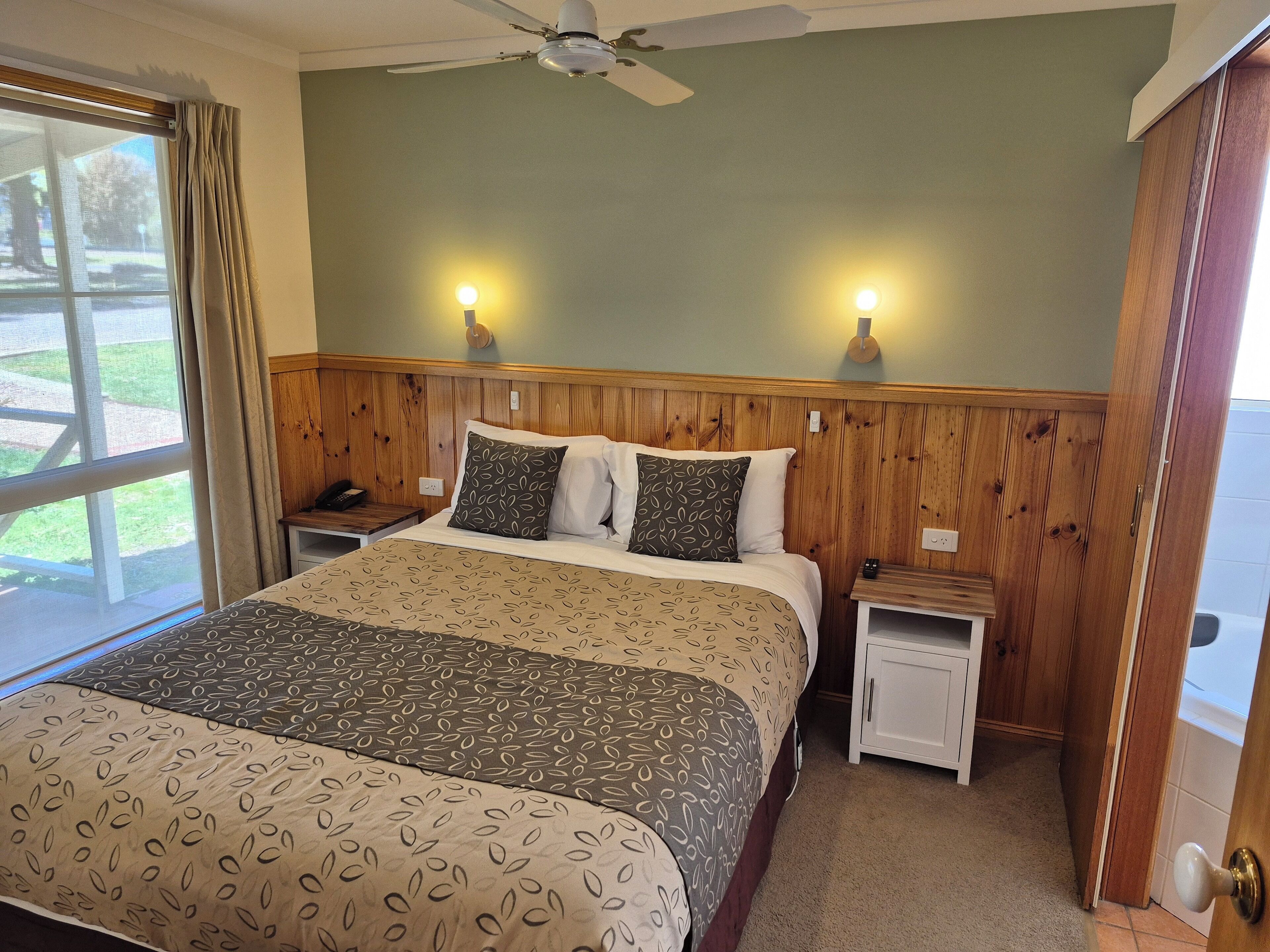 Habitación de hotel con cama, cabecera de madera, dos mesitas de noche, una mesita auxiliar, ventilador de techo y vistas al exterior a través de una ventana.