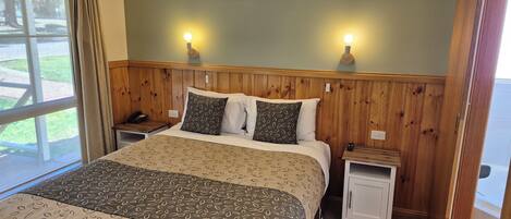 Habitación de hotel con cama, cabecera de madera, dos mesitas de noche, una mesita auxiliar, ventilador de techo y vistas al exterior a través de una ventana.
