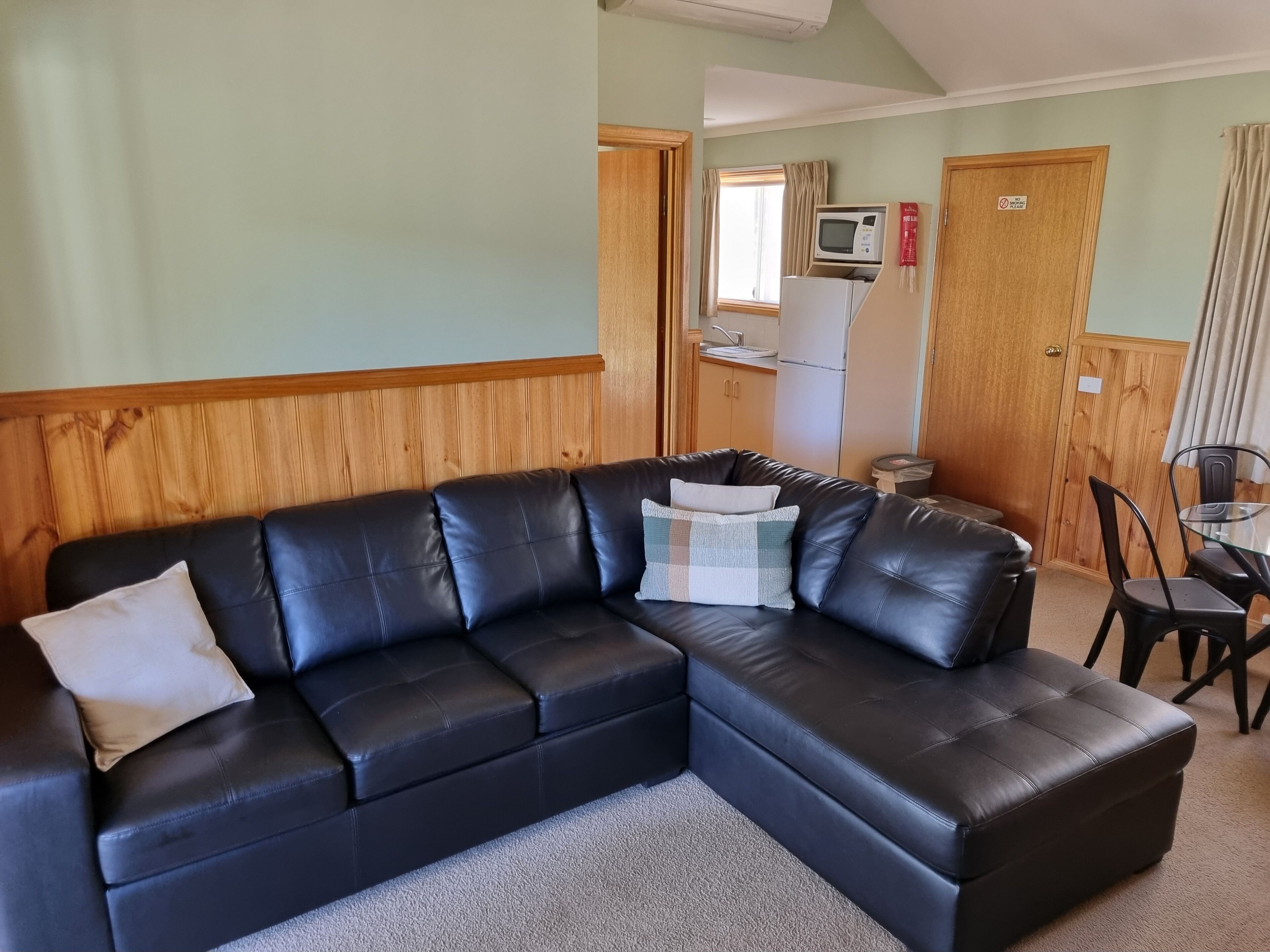 Uma sala de estar com um sofá de couro preto, uma mesinha de vidro e uma cozinha com micro-ondas e geladeira.
