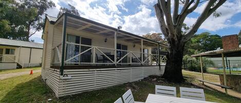 A white mobile home with a covered porch, a large tree in the foreground, and a grassy yard.