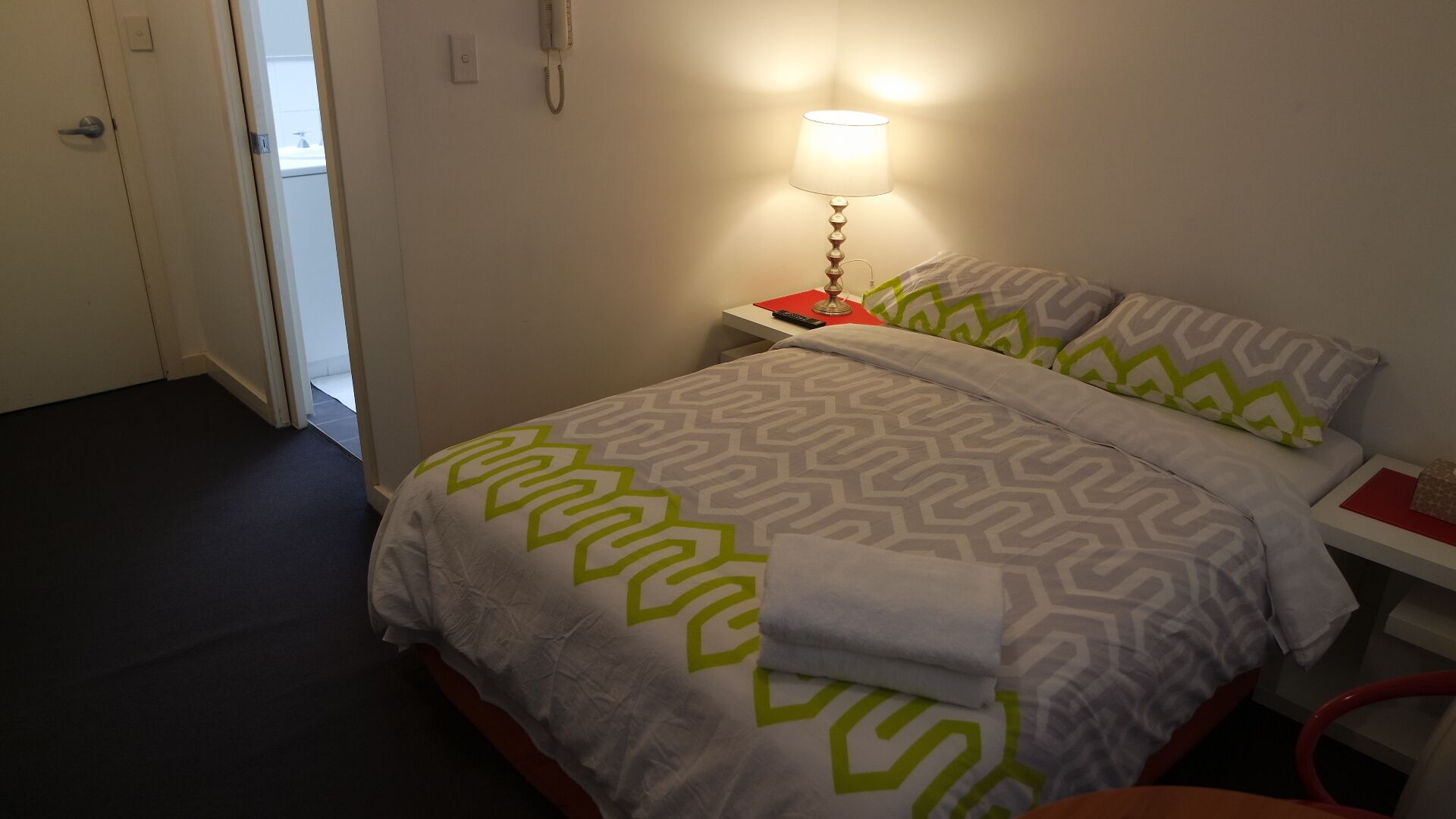 A neatly made bed with a patterned bedspread, a bedside lamp, and a nightstand with a phone and remote control.