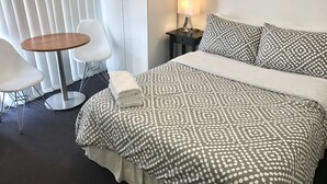 A hotel room with a neatly made bed, a small round table, two white chairs, a nightstand with a lamp, and a window with blinds. - Cityview Studio Accommodation (Leichhardt)