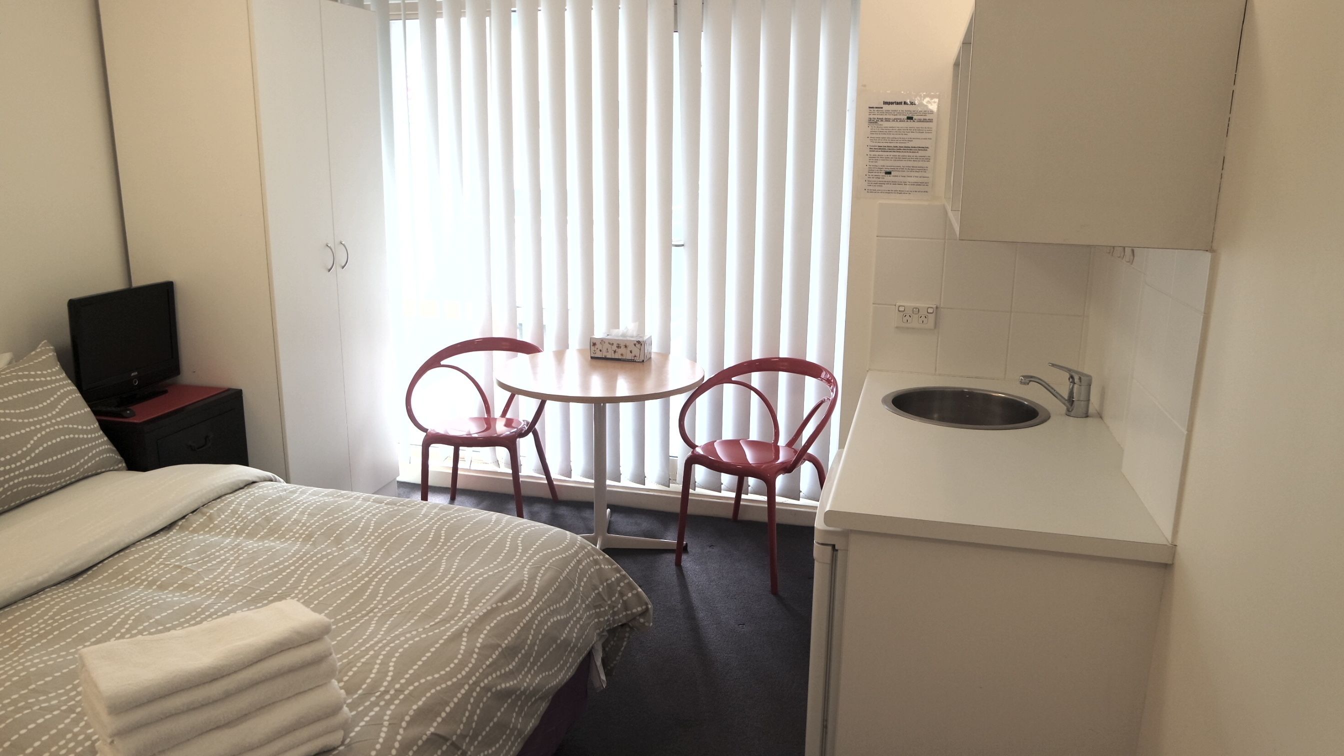Une petite chambre d’hôtel comprenant un lit, une télévision, un bureau avec une chaise, une fenêtre avec des stores et un lavabo.