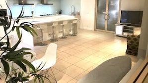 A modern kitchen with a central island, bar stools, a dining area with a white table and chairs, a grey sofa, and a television in a cabinet. - Cityview Studio Accommodation (Leichhardt)
