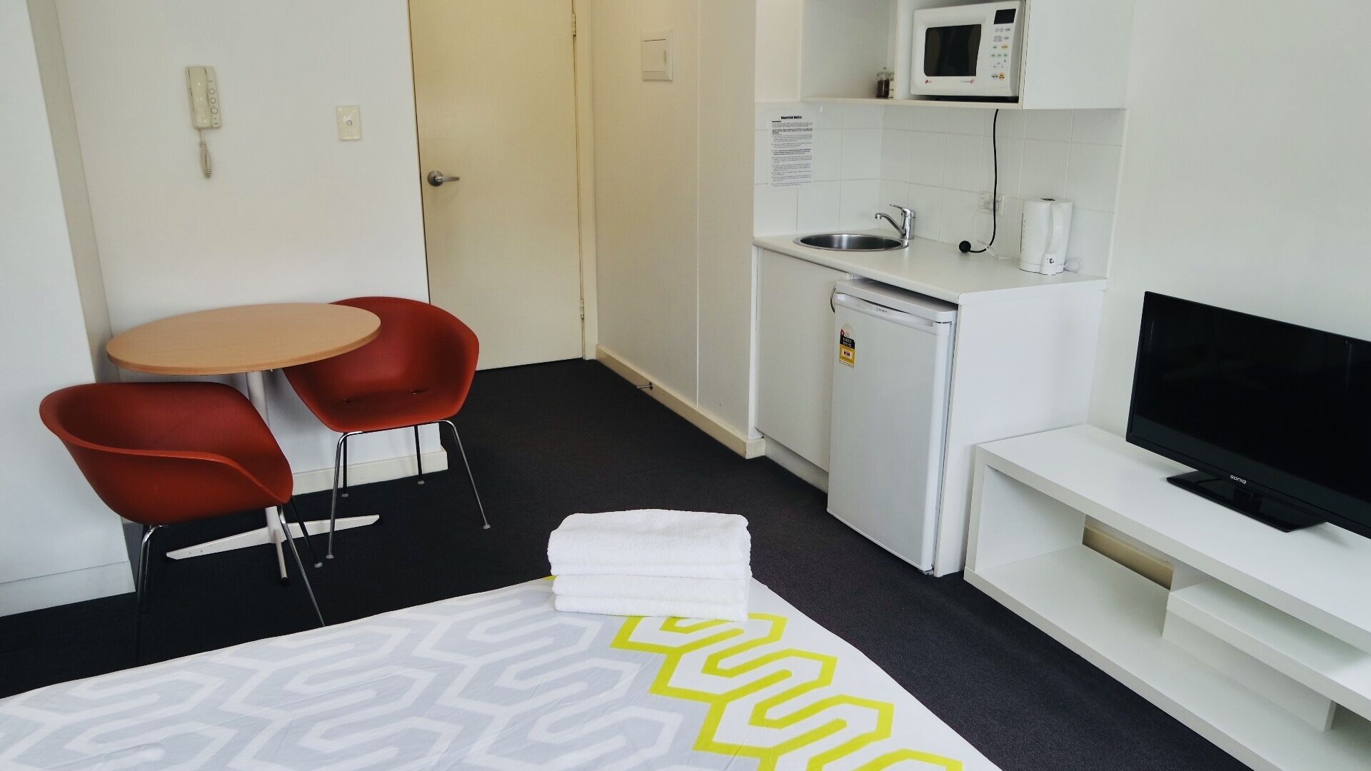 A compact hotel room with a bed, a small kitchenette, a TV, and two red chairs.