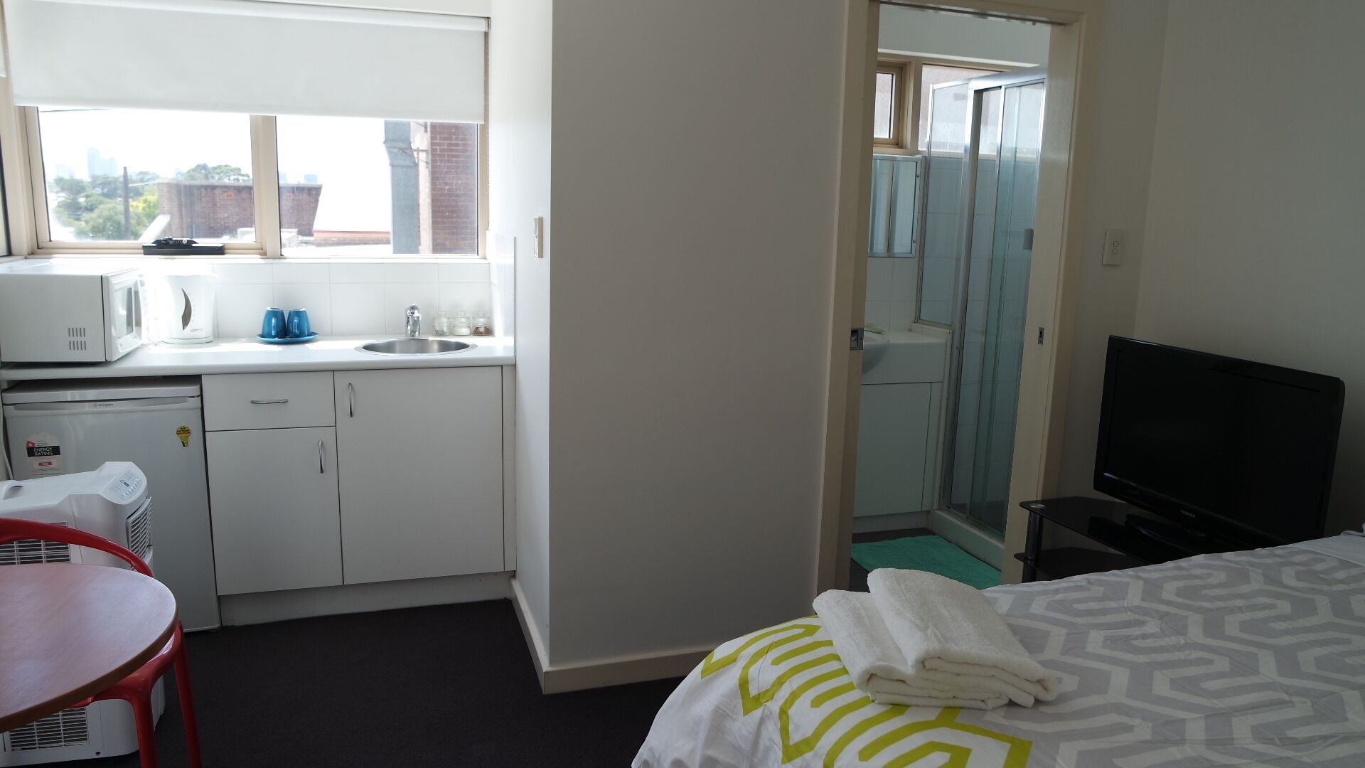 A hotel room with a kitchenette, a bed with a patterned bedspread, a TV, and a bathroom with a glass shower enclosure.