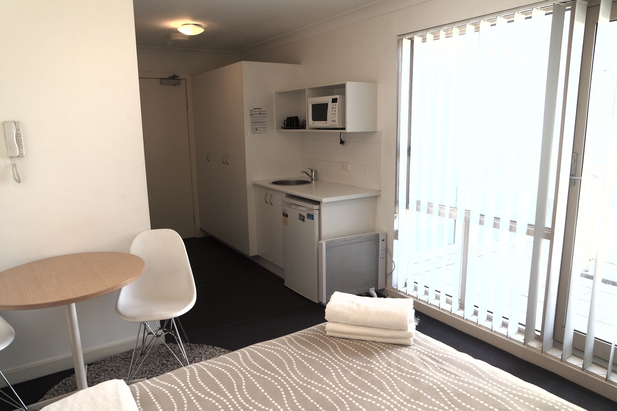 Una habitación de hotel pequeña y funcional con una cama individual, una mesita, una silla, una kitchenette con microondas y vistas al exterior a través de un ventanal grande.