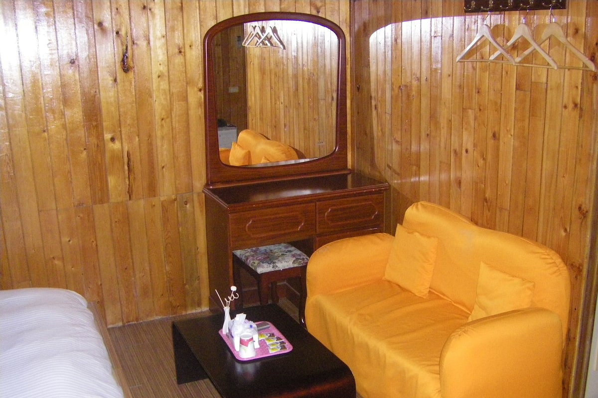 Una habitación con paneles de madera, un aparador de madera con espejo, un sofá naranja, una mesita con una bandeja rosa y una cama con ropa de cama blanca.