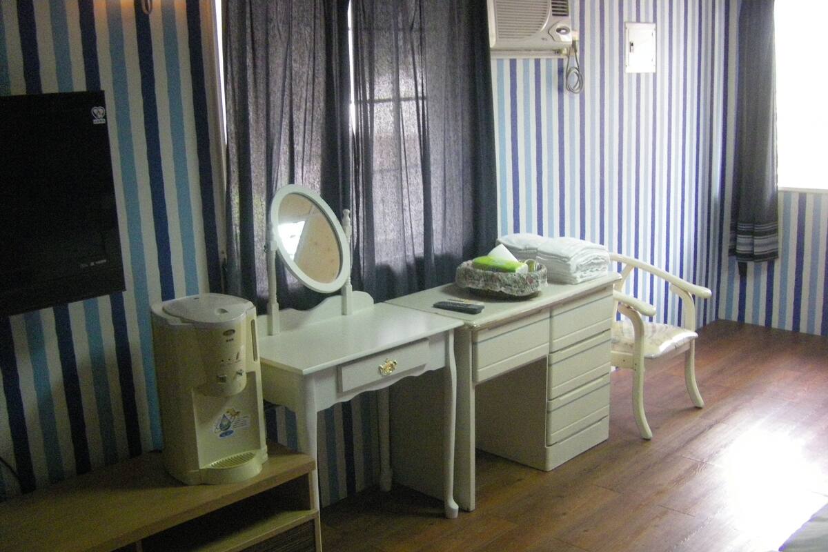 Una habitación con tocador blanco, espejo, silla y una mesita con secador de pelo.