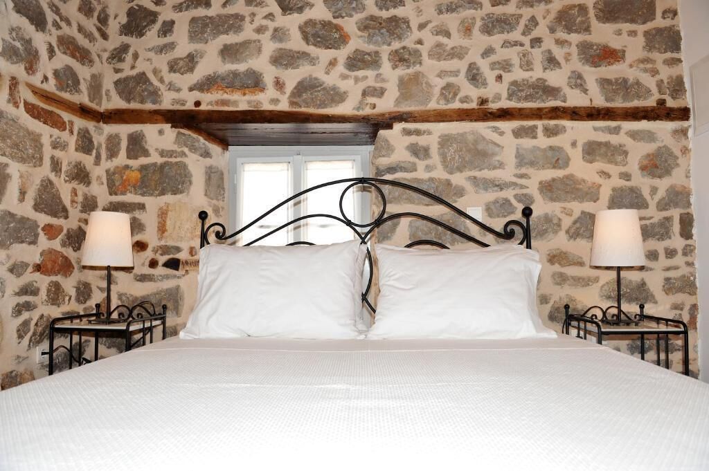 Un dormitorio con pared de piedra, una cama con ropa blanca, dos mesitas de noche con lámparas y una ventana sobre la cama.