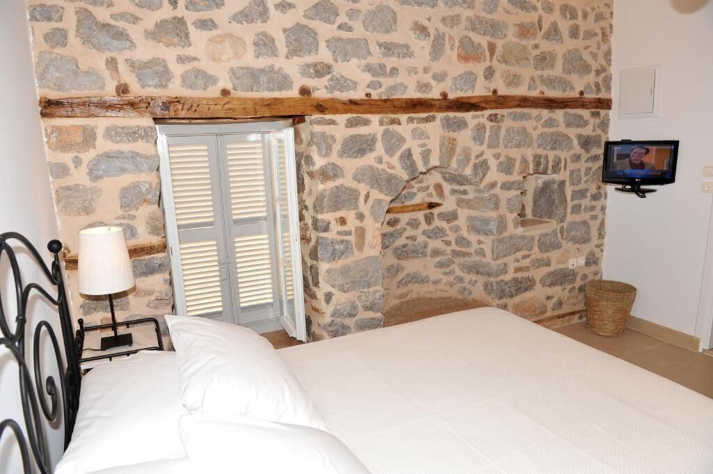A bedroom with a stone wall, a bed with white bedding, a wooden headboard, a bedside lamp, a television, and a wicker basket.