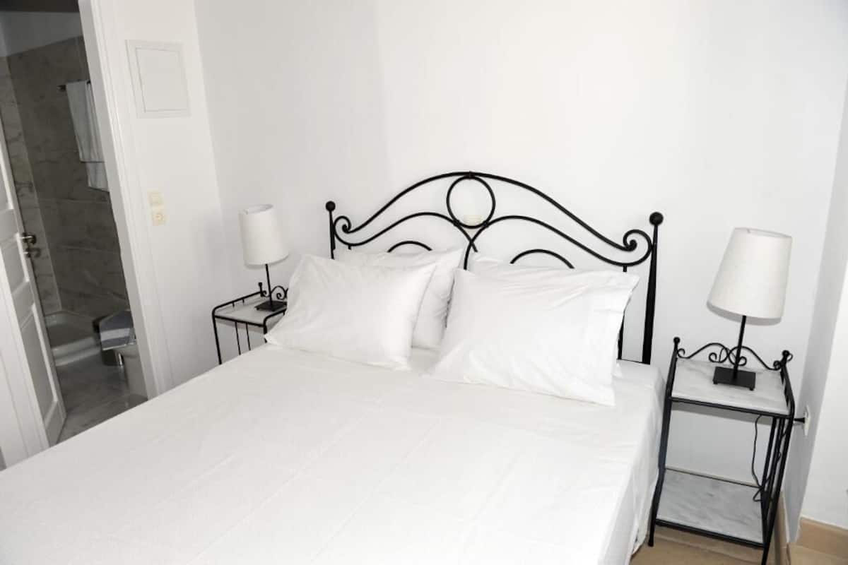 A neatly made bed with white linens in a room with white walls and a black metal headboard. There are two bedside tables with lamps on either side of the bed.