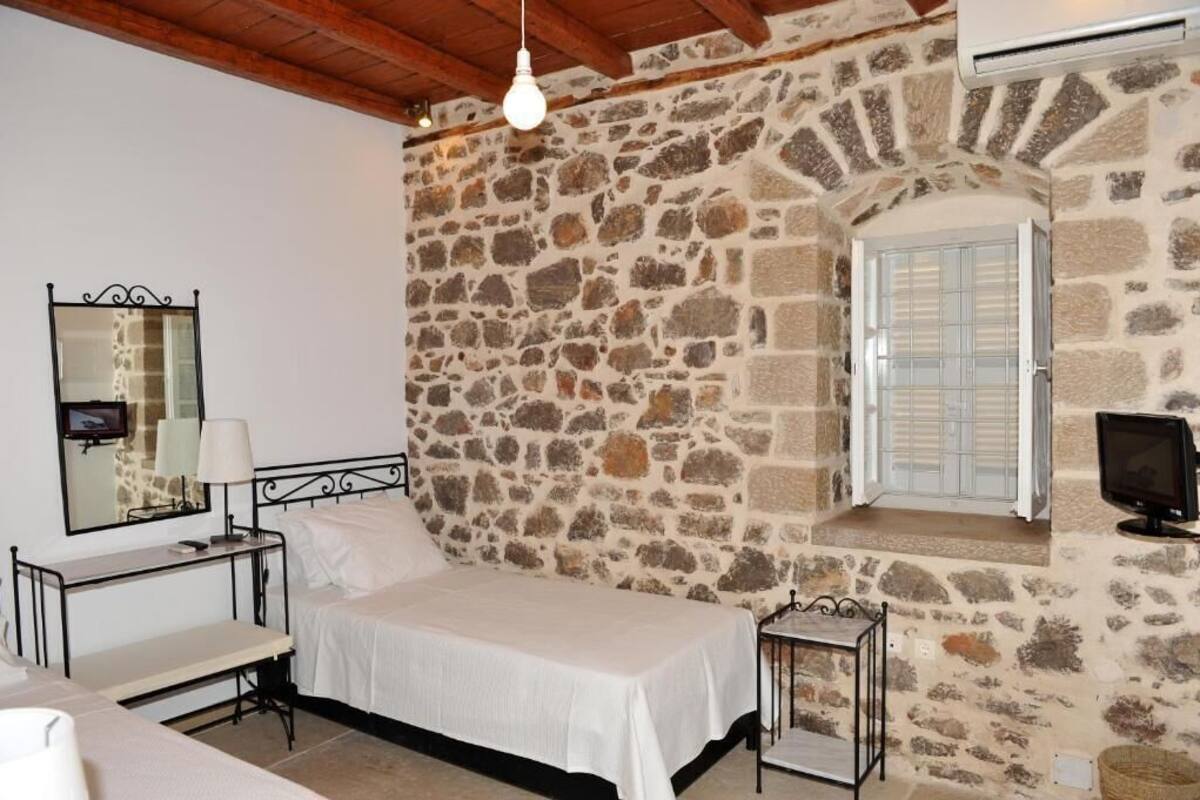 A bedroom with a stone wall, a bed, a bedside table with a lamp, a window with shutters, and a television.
