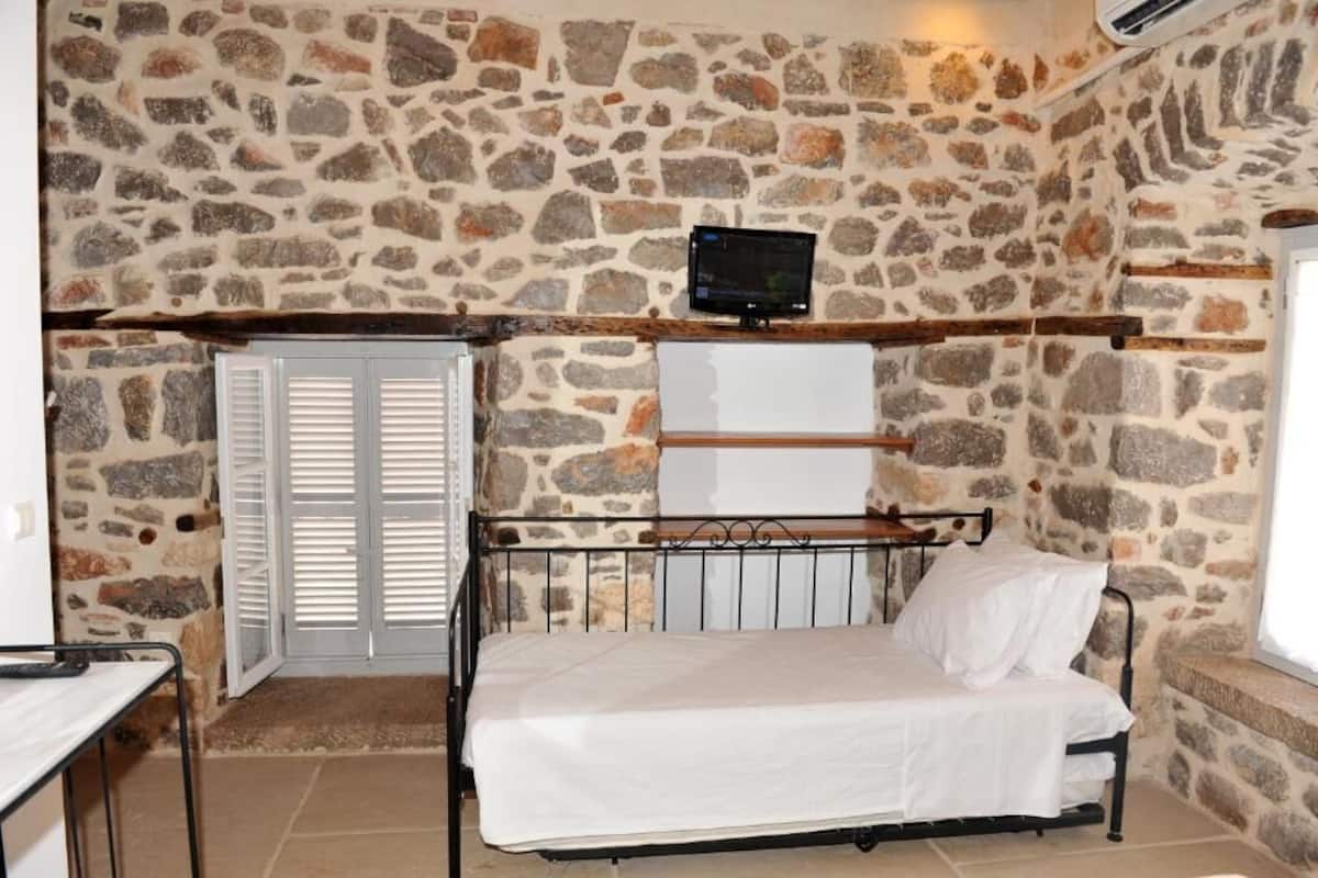 A stone-walled room with a bed, a television mounted on the wall, and a window.