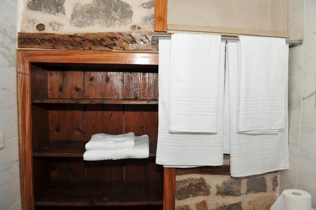A bathroom with a wooden shelf holding folded towels, a stone wall, and a tiled floor.