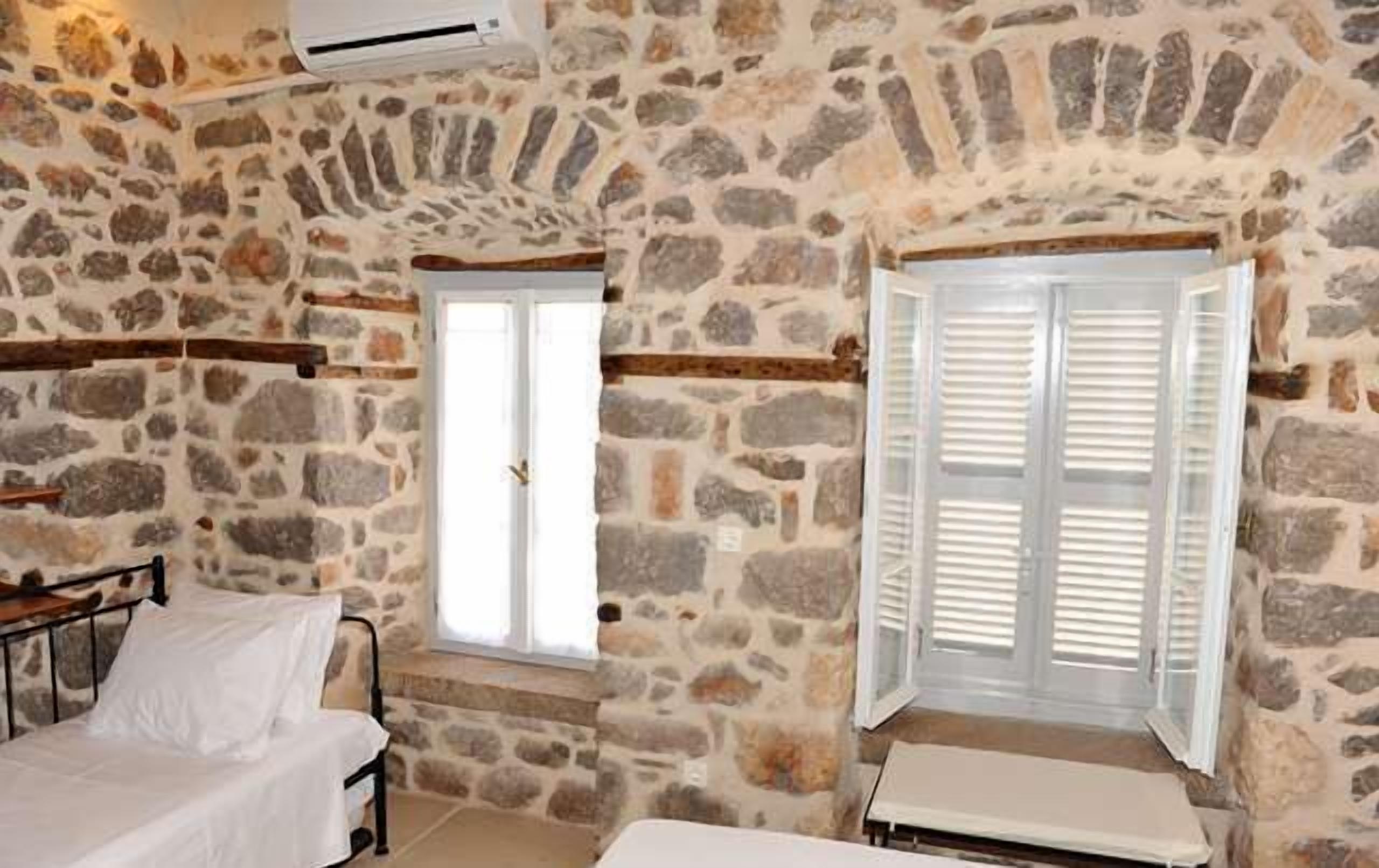 A stone-walled room with a bed, a window with shutters, and an air conditioning unit.