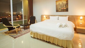 A hotel room with a large bed, two bedside lamps, a chair, a small table, and a view of the city at night. - Soontree House (Udon Thani)