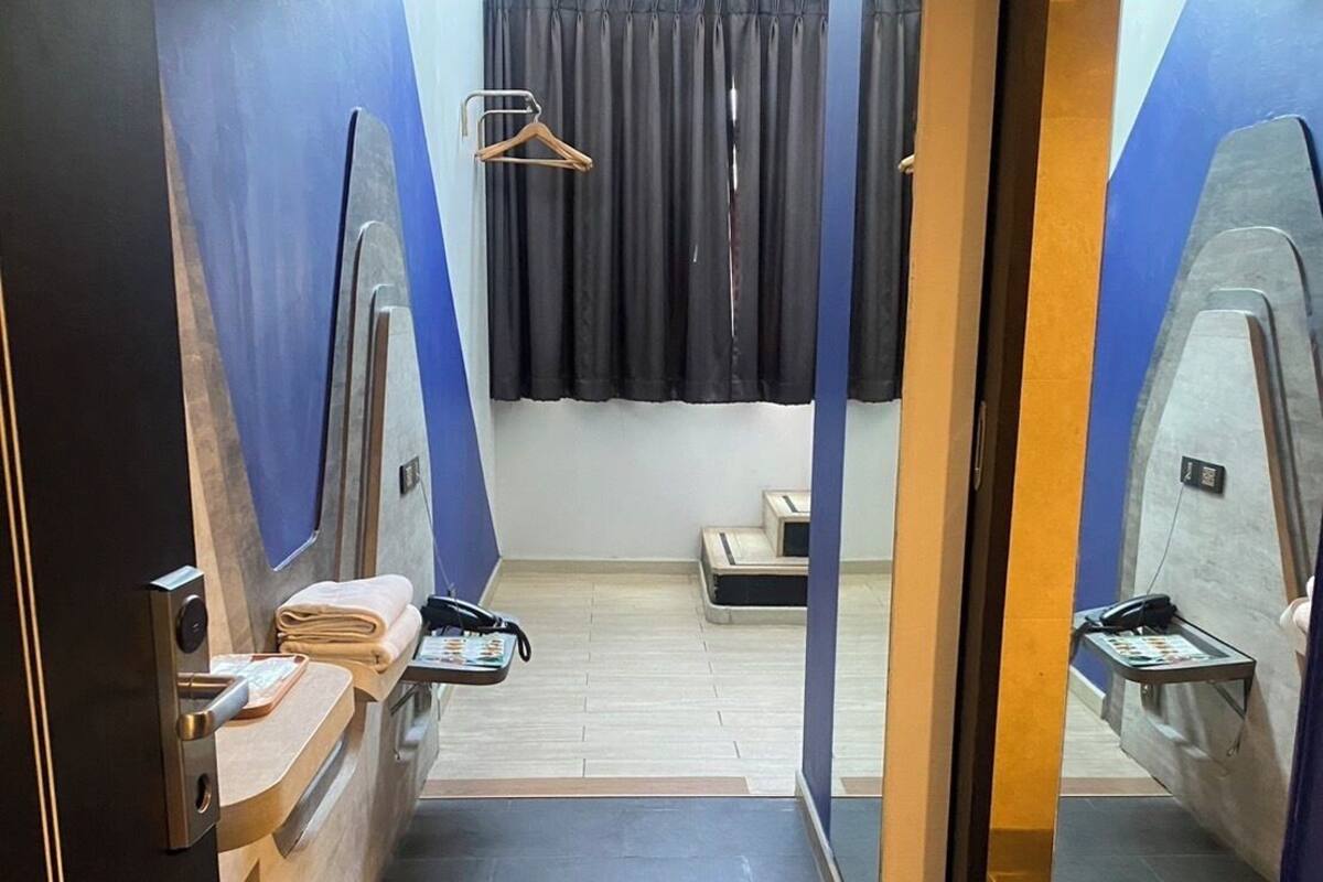 A narrow hallway leading to a room with a bed, a small table, and a chair. There is a window with curtains and a door on the right side.