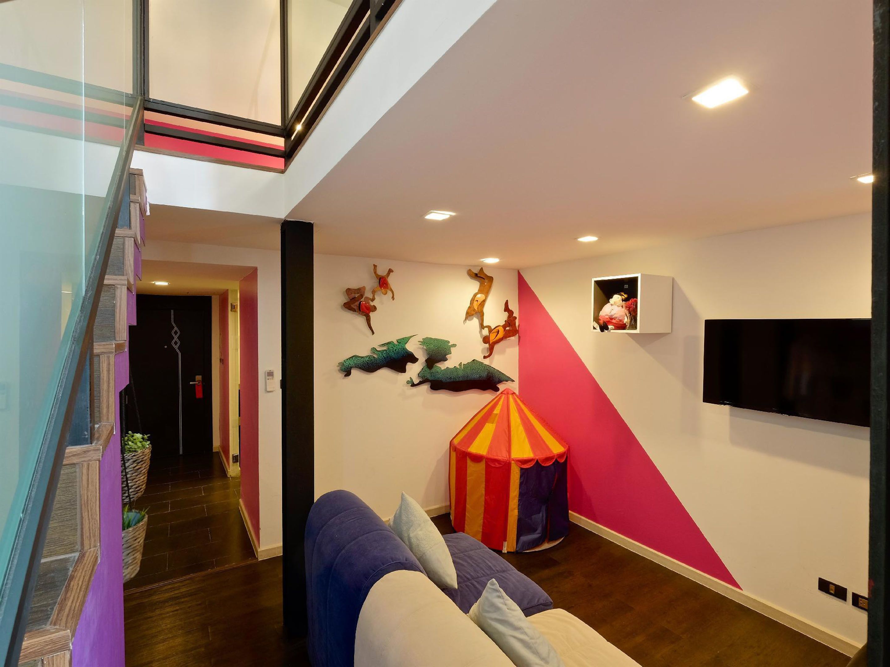 Ein modernes Wohnzimmer mit einer Glastreppe, einer farbenfrohen Wandkunstinstallation, einem an der Wand montierten Fernseher und einem komfortablen Sofa.