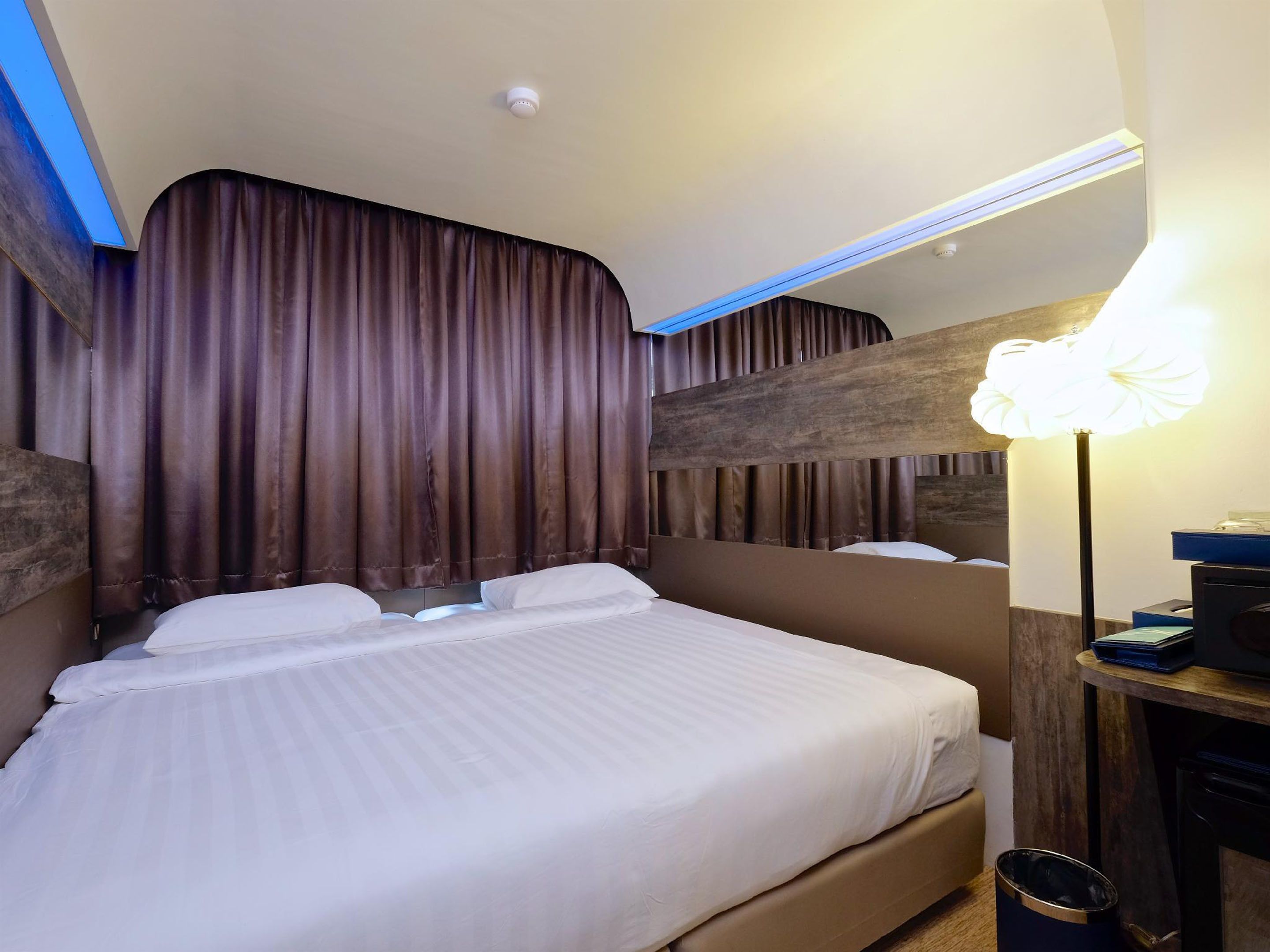 A hotel room with a large bed, dark curtains, a modern headboard, a bedside table with a lamp, and a trash bin.