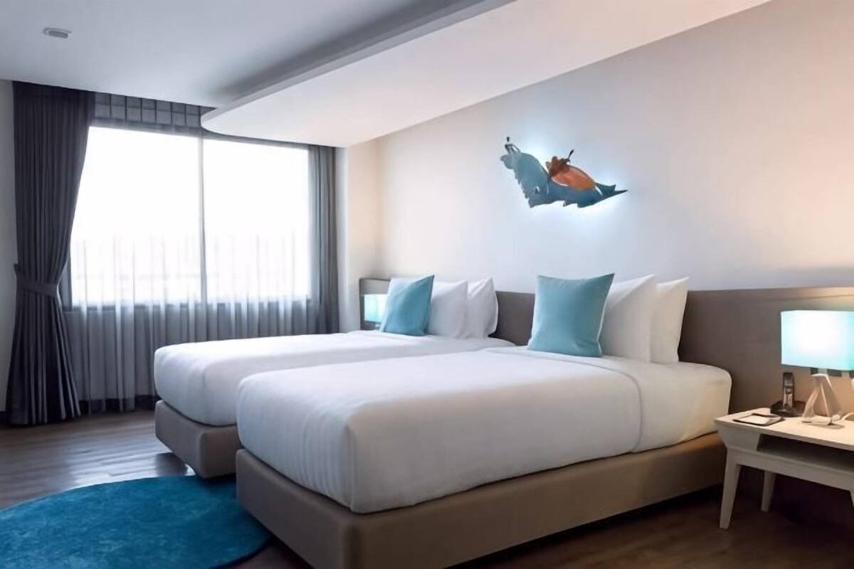 A hotel room with two beds, a nightstand, a lamp, and a window with curtains.