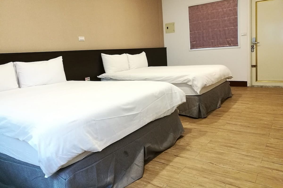 A hotel room with two single beds, a wooden floor, a beige wall, and a window with a blind.