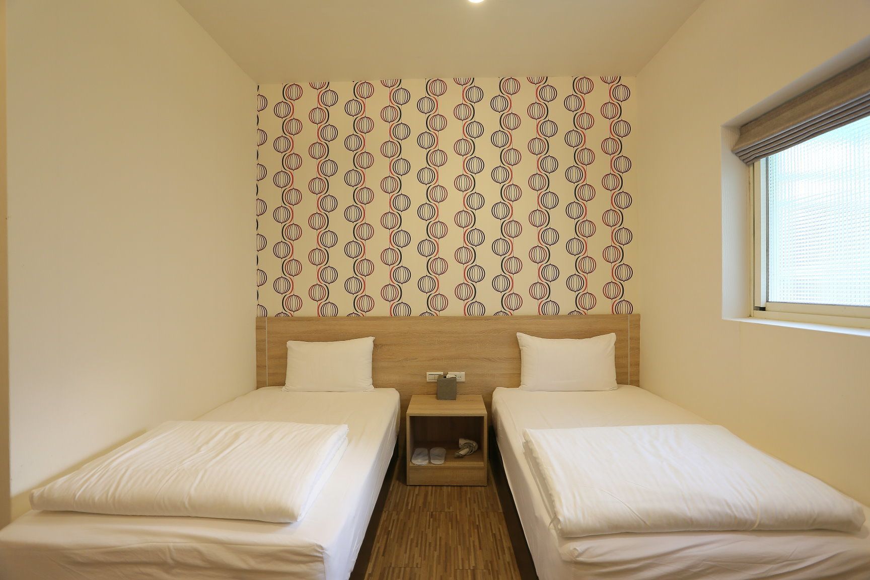 A double room with two beds, a wooden headboard, a small bedside table, and a window with blinds.