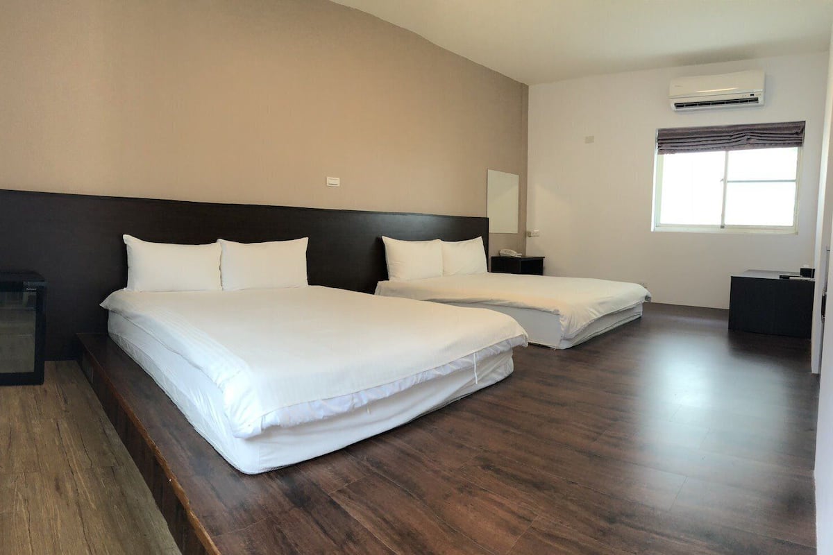 A hotel room with two beds, a wooden floor, a large mirror, an air conditioning unit, and a window with curtains.