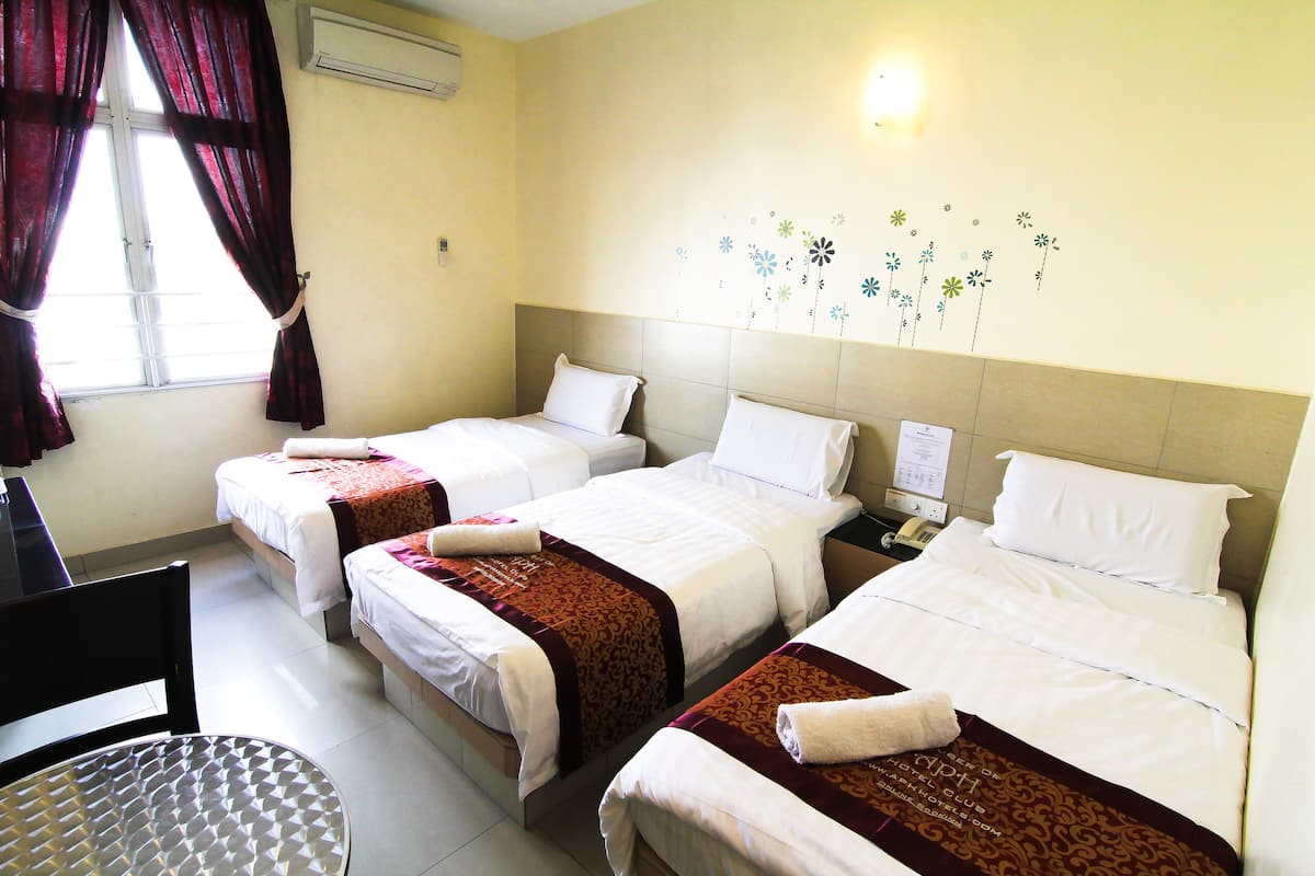 A hotel room with three single beds, each with white linens and brown decorative borders. There is a desk with a chair, a window with red curtains, and a wall-mounted air conditioning unit.